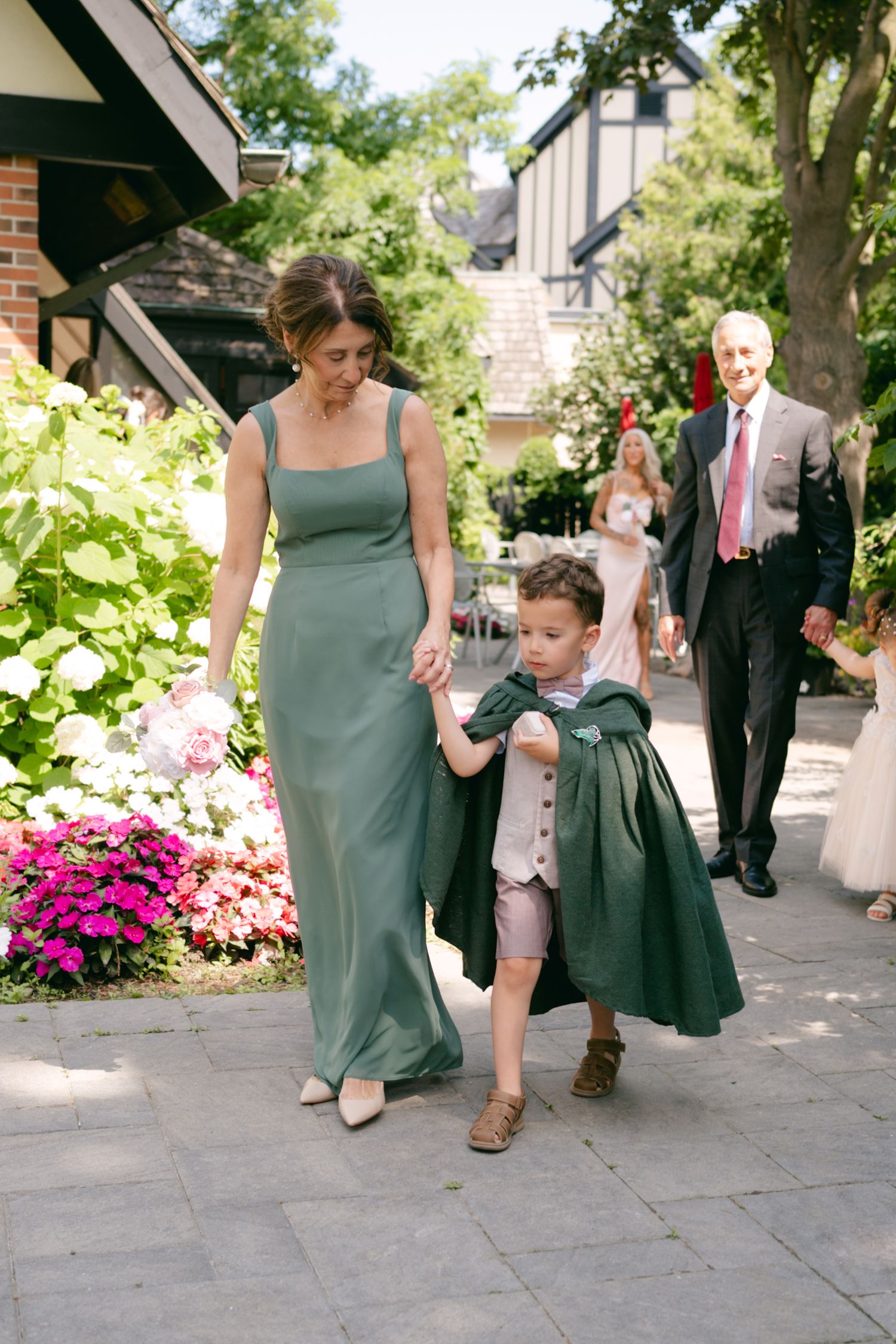 Wedding party members in green dresses and suits walk along a garden path during an outdoor celebration.