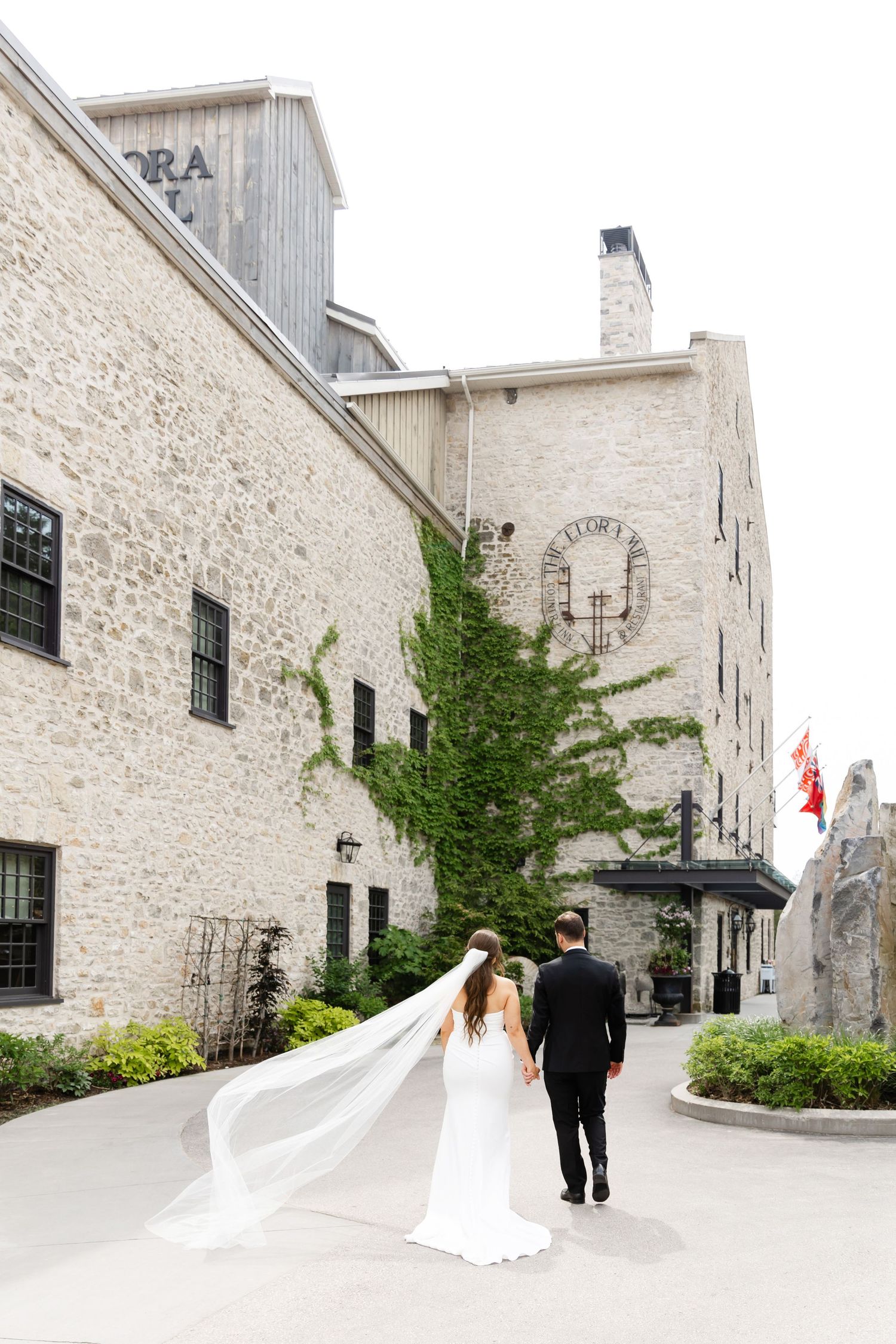 Bride and groom walk hand in hand past an ivy-covered stone building on their wedding day.