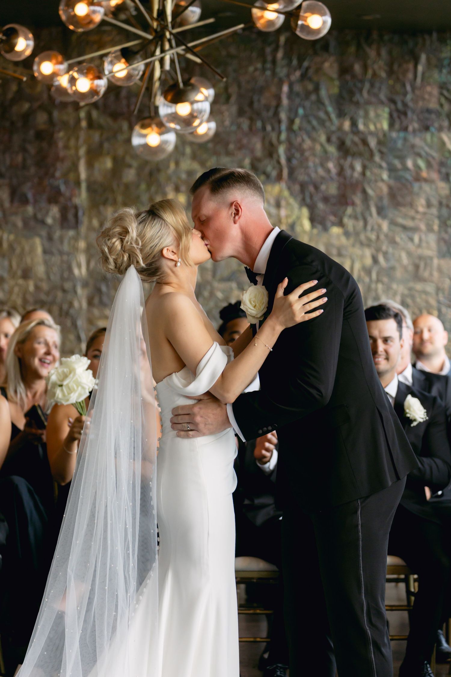 Joyful newlyweds share first kiss as wedding guests cheer beneath a modern globe chandelier.