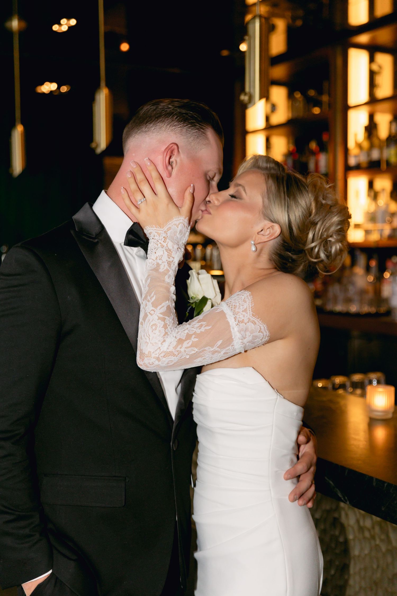 Bride in lace-sleeved white gown kisses groom in black tuxedo at a warmly lit bar during wedding reception.