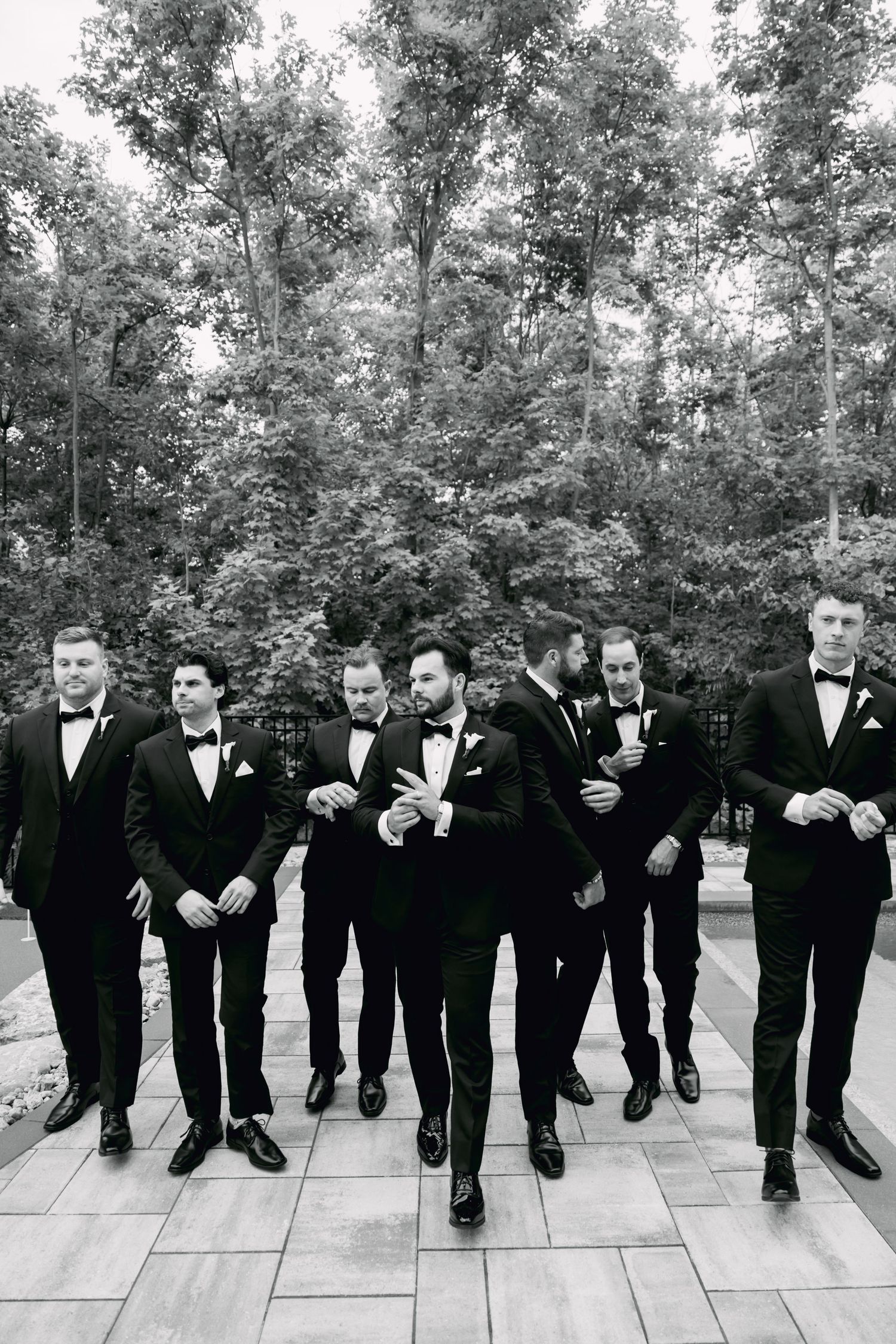 Groomsmen in matching black tuxedos and bow ties walk confidently together in black and white wedding photo.