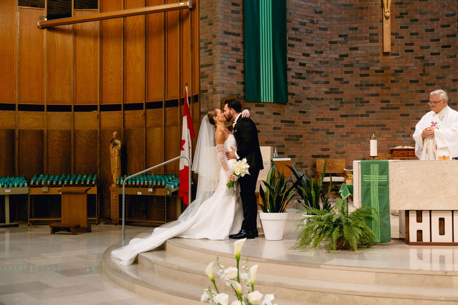 Bride and groom share first kiss at church altar during wedding ceremony with priest officiating.