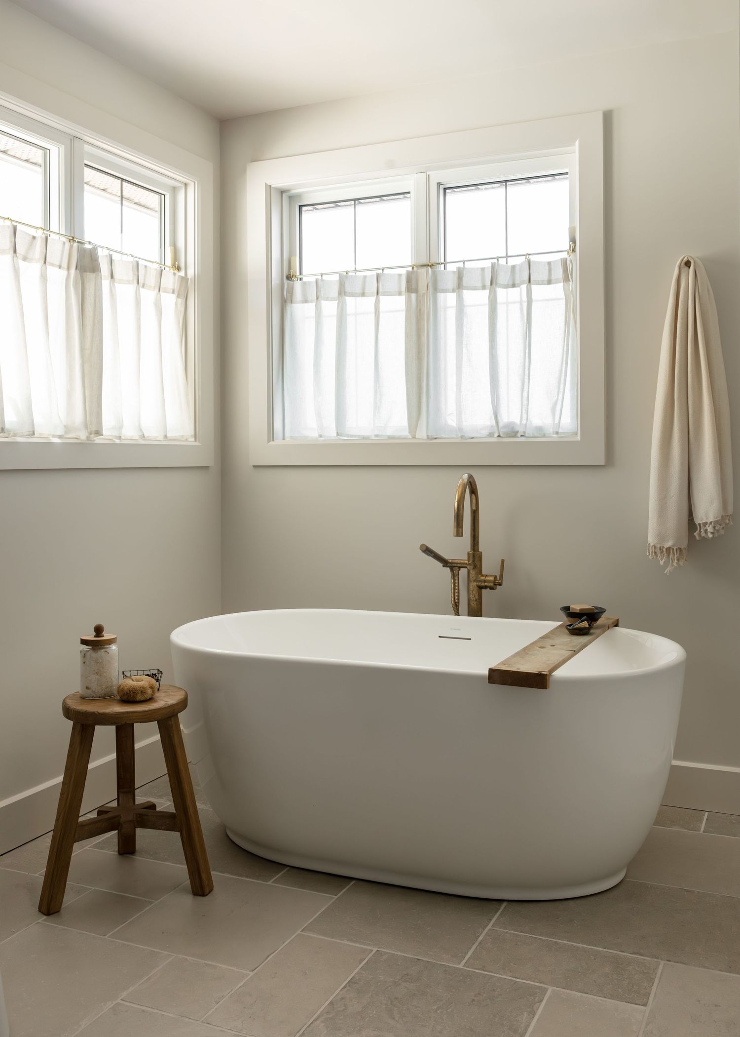 Luxurious freestanding white bathtub with wooden stool and brass fixtures in a bright bathroom.