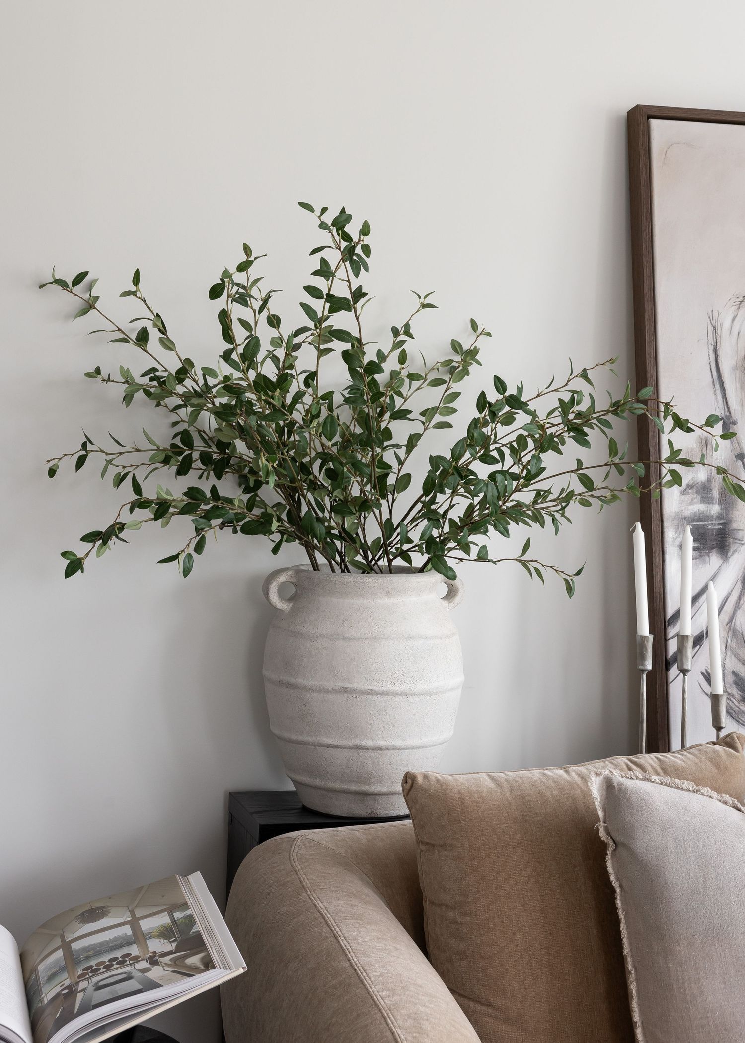 Large white ceramic vase with fresh eucalyptus branches next to framed artwork and beige sofa.