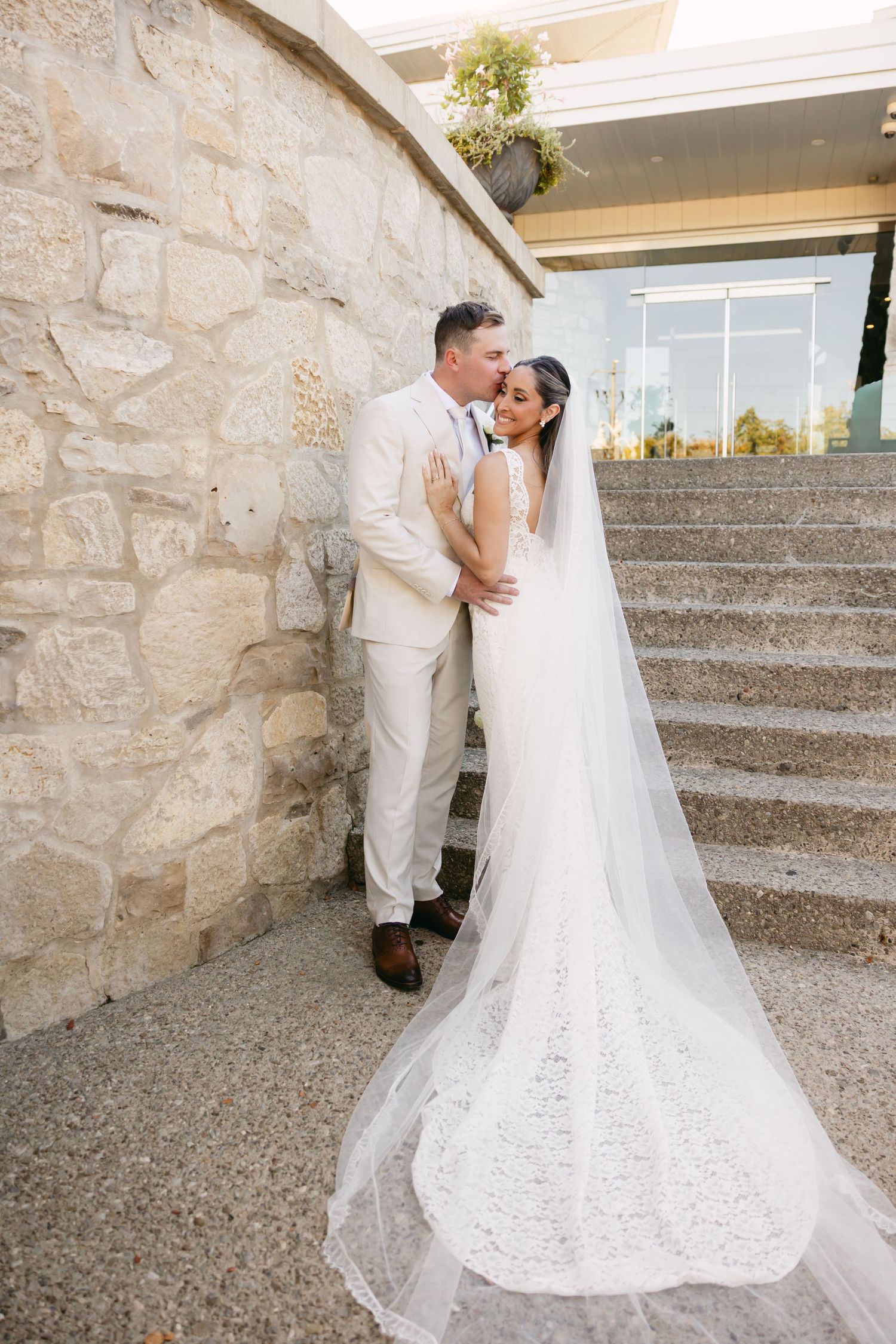 A wedding couple embraces on stone steps with a flowing white veil trailing behind them.