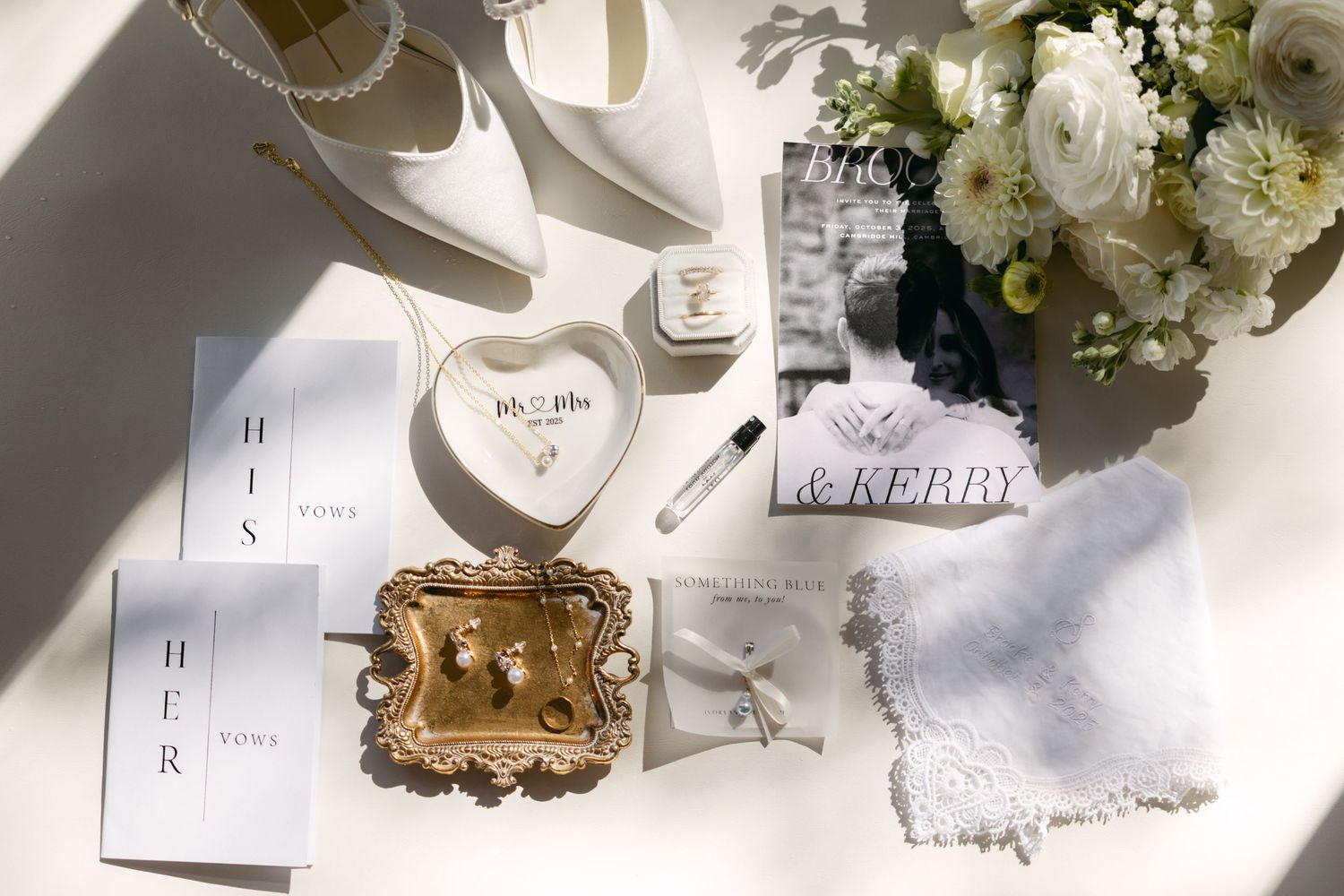Wedding details flatlay with white flowers, lace handkerchief, and gold accents on white surface.