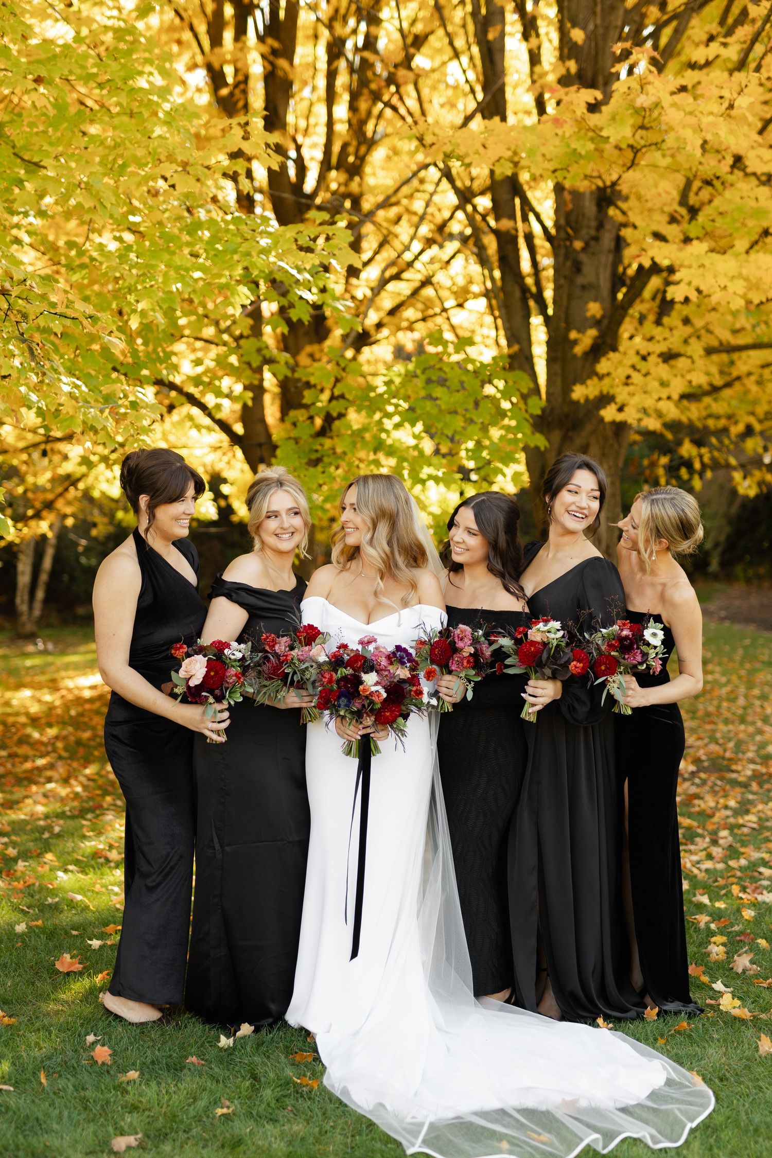 Bridesmaids in black dresses hold burgundy bouquets while posing together on grass with fall foliage in background.