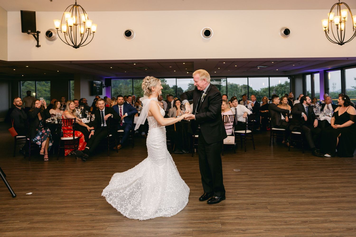 Wedding guests dancing and celebrating on a wooden dance floor at a reception venue with elegant chandeliers.