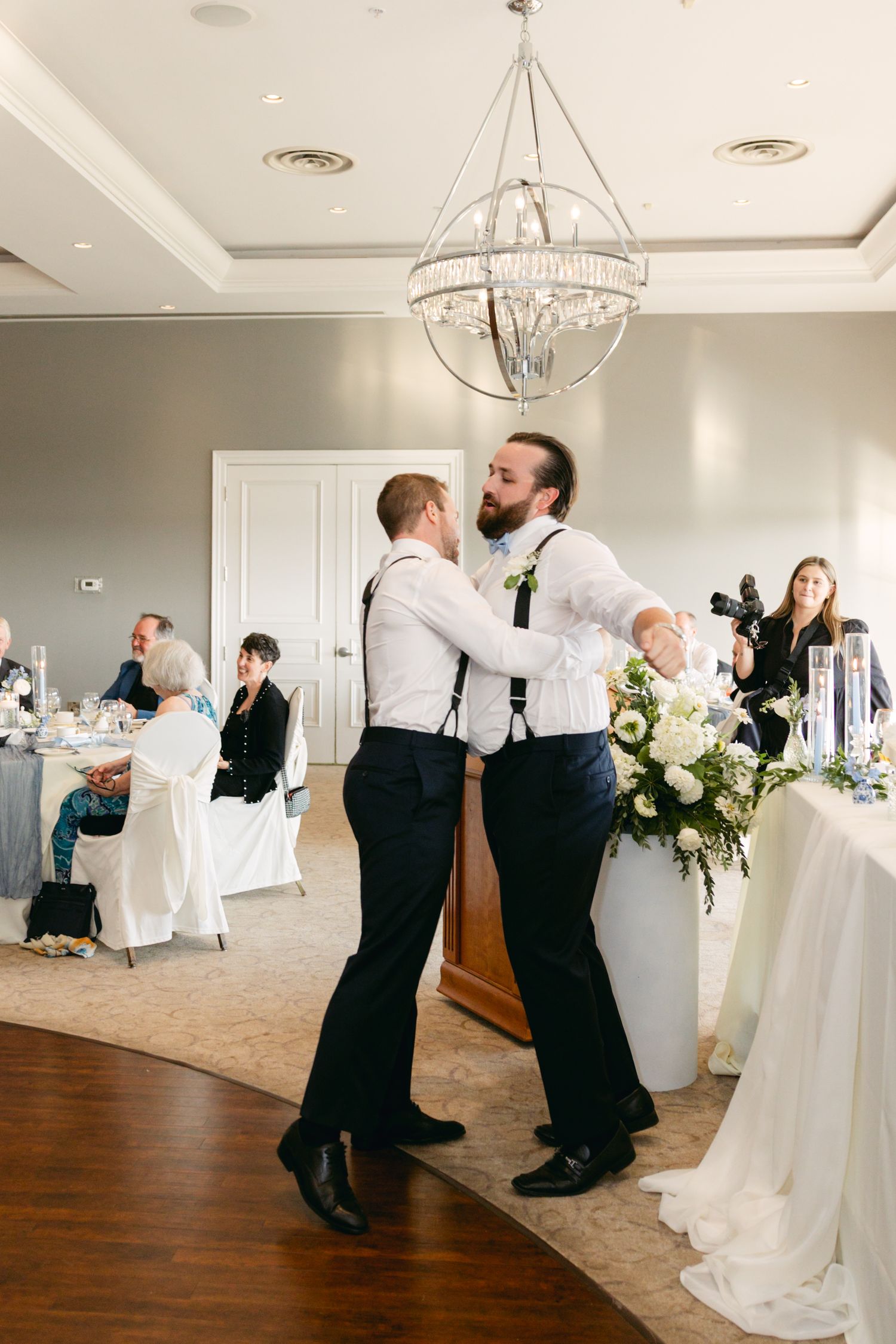 Wedding party members executing synchronized dance moves on hardwood floor under crystal chandeliers.