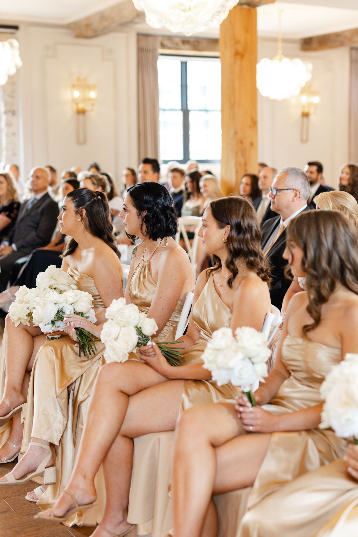 Bridesmaids in gold satin dresses hold white bouquets while seated at indoor wedding ceremony.