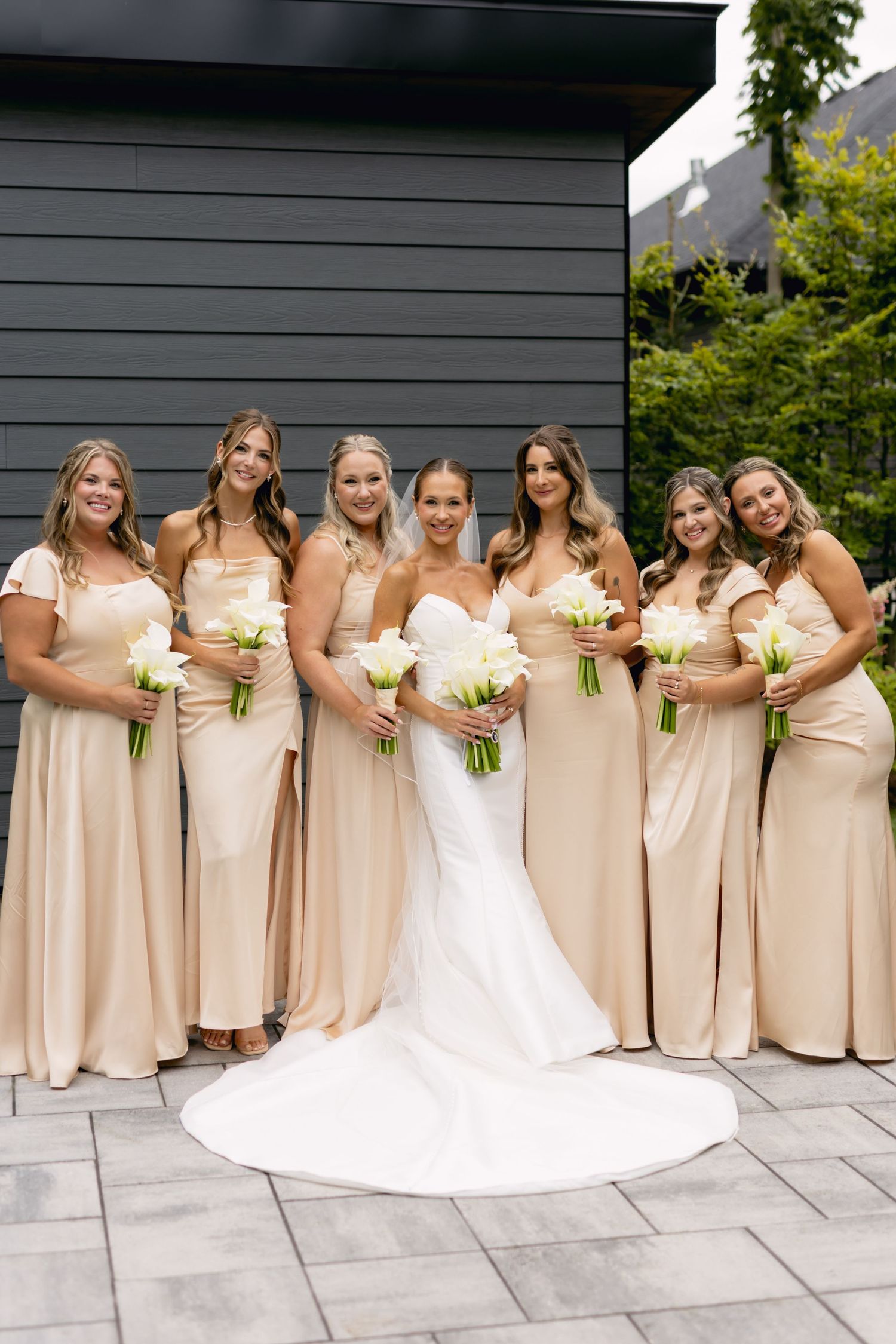 Bride with six bridesmaids in champagne gowns holding white calla lily bouquets posing outdoors.