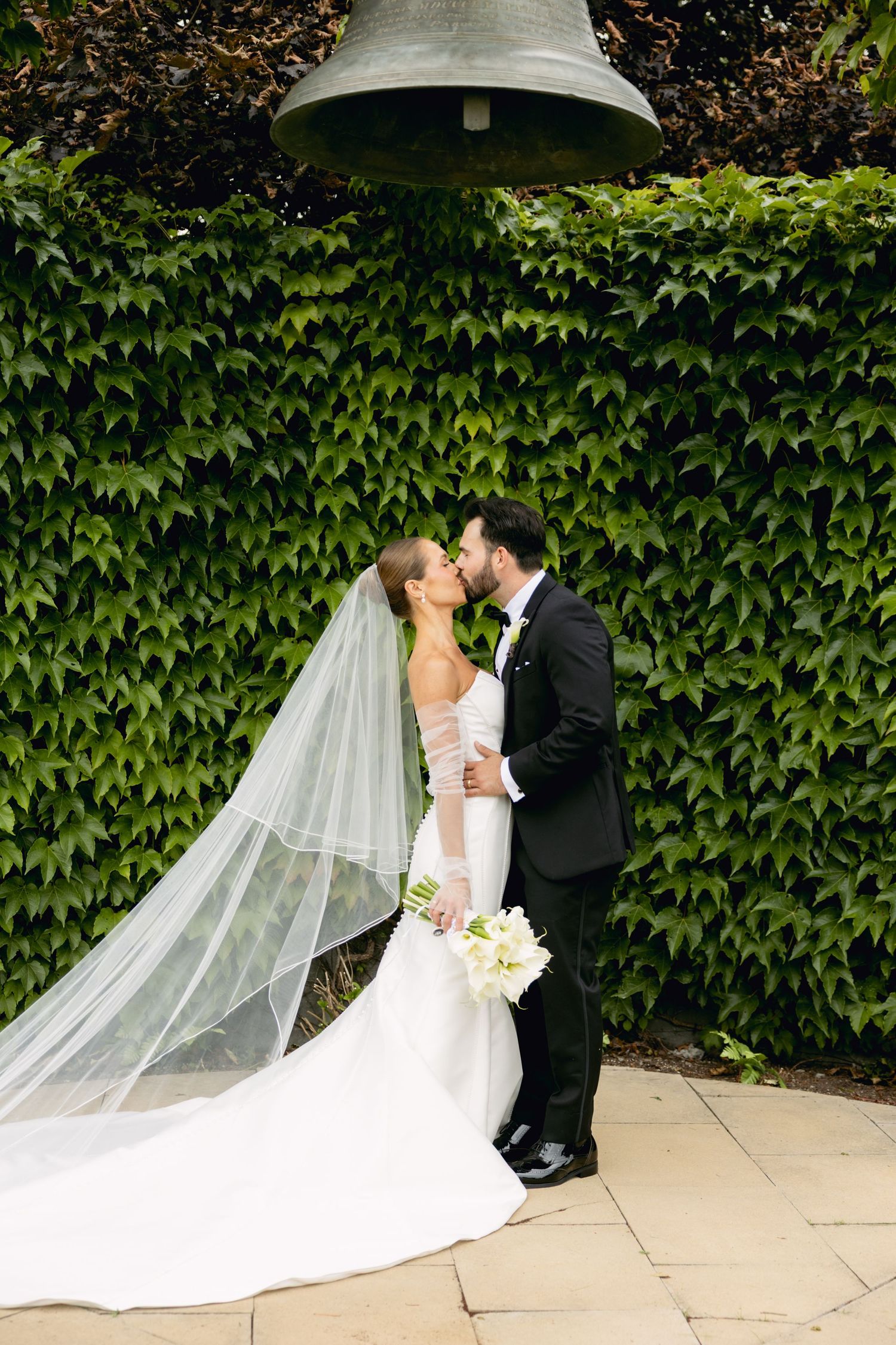 Bride and groom kiss under large bell surrounded by lush green ivy wall.