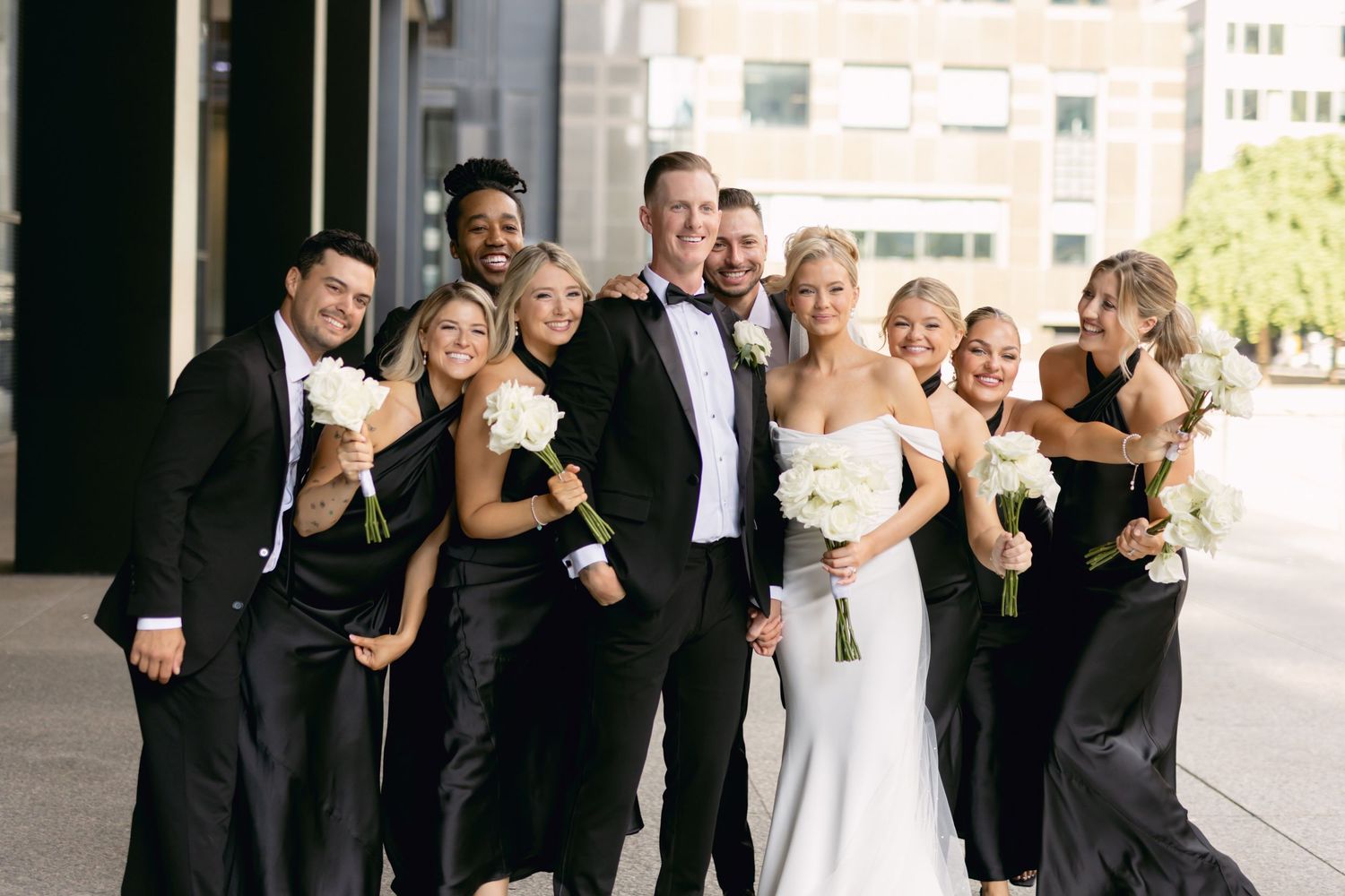Wedding party in black attire with bride in white gown posing outdoors holding white bouquets in urban setting.