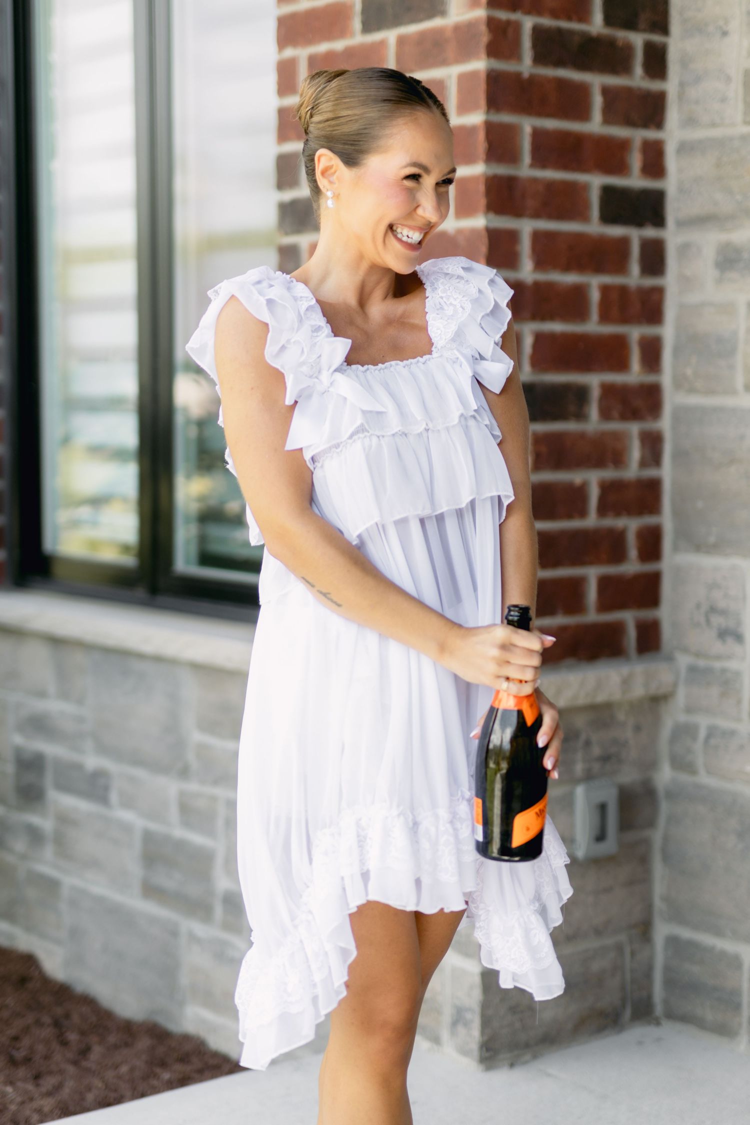 Smiling bride in white ruffled dress holding Veuve Clicquot champagne bottle outside brick building.
