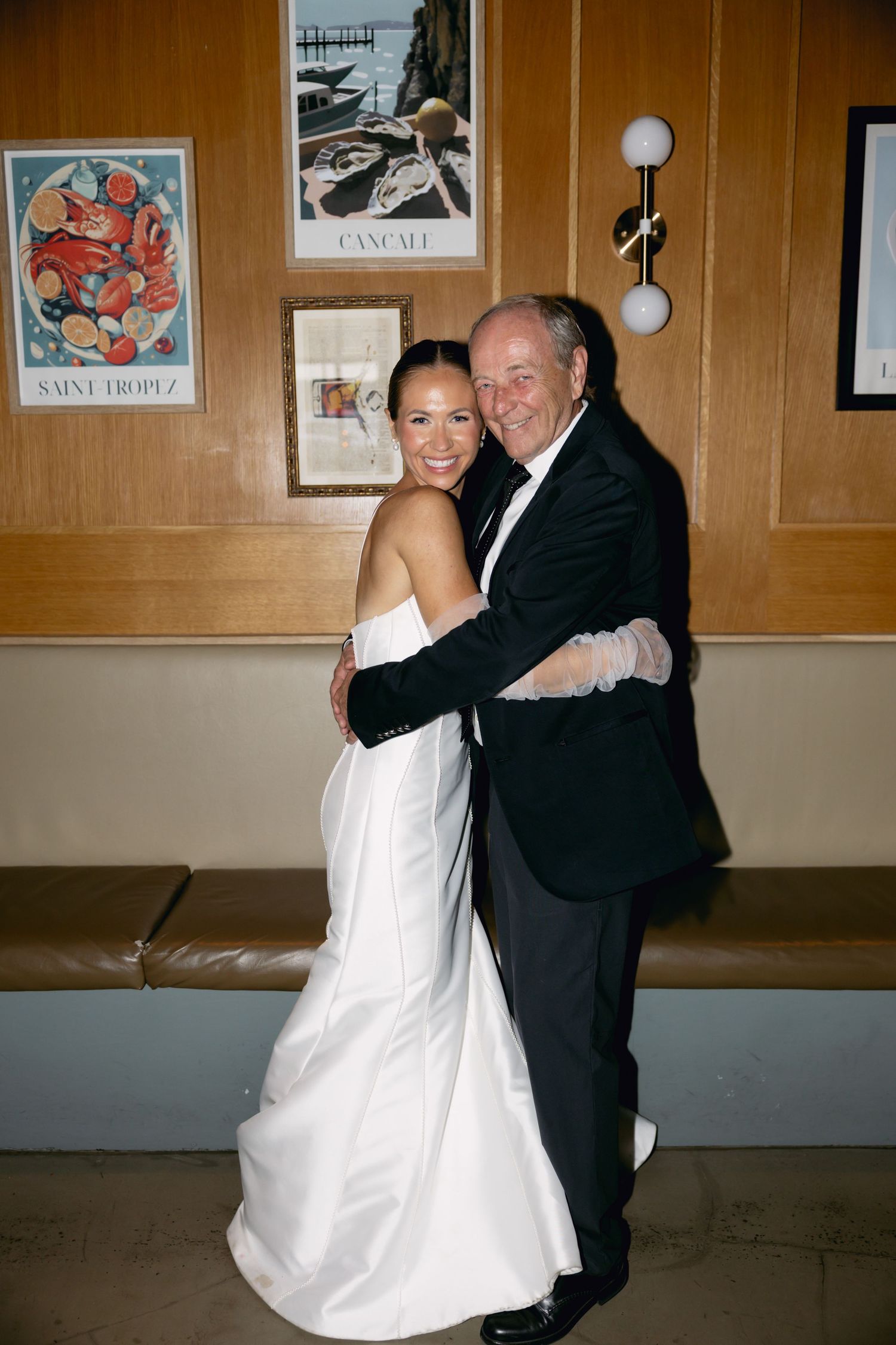 Bride in white strapless gown shares a warm embrace with older gentleman in black tuxedo at wedding reception.