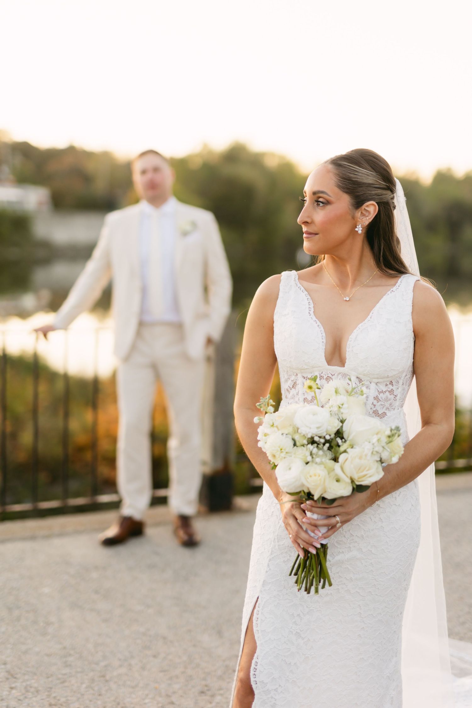 Elegant bride in lace dress holding white floral bouquet against sunset backdrop.
