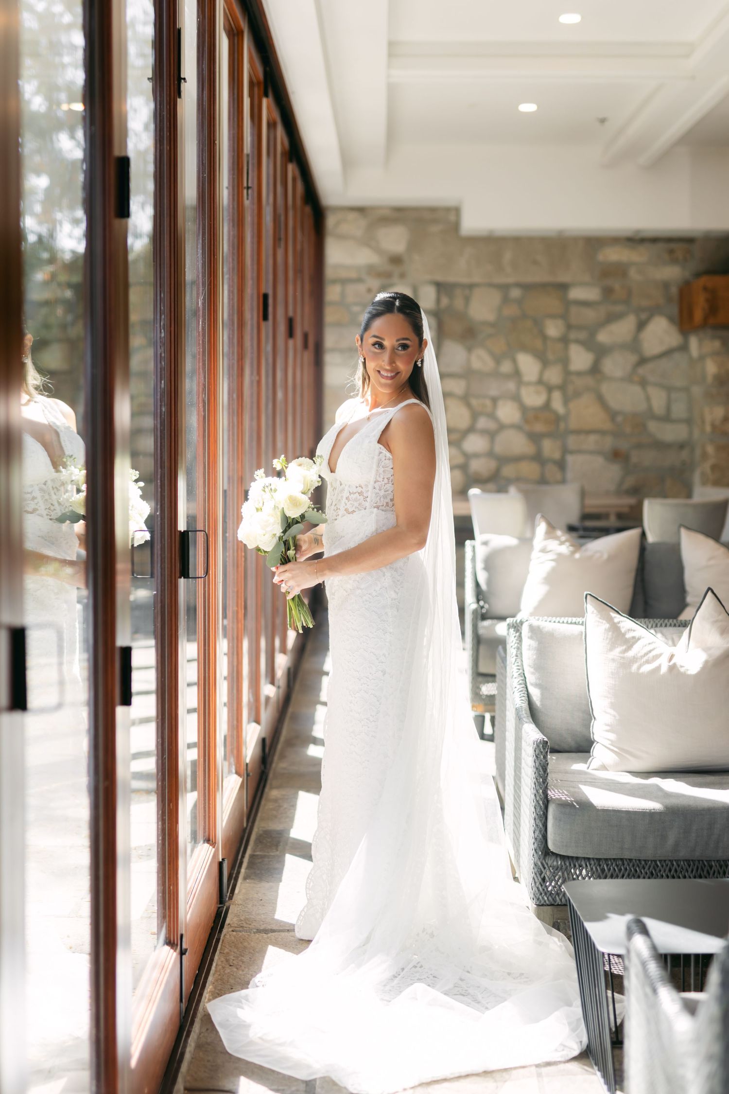A bride holds a white bouquet while standing in a sunlit room with stone walls.