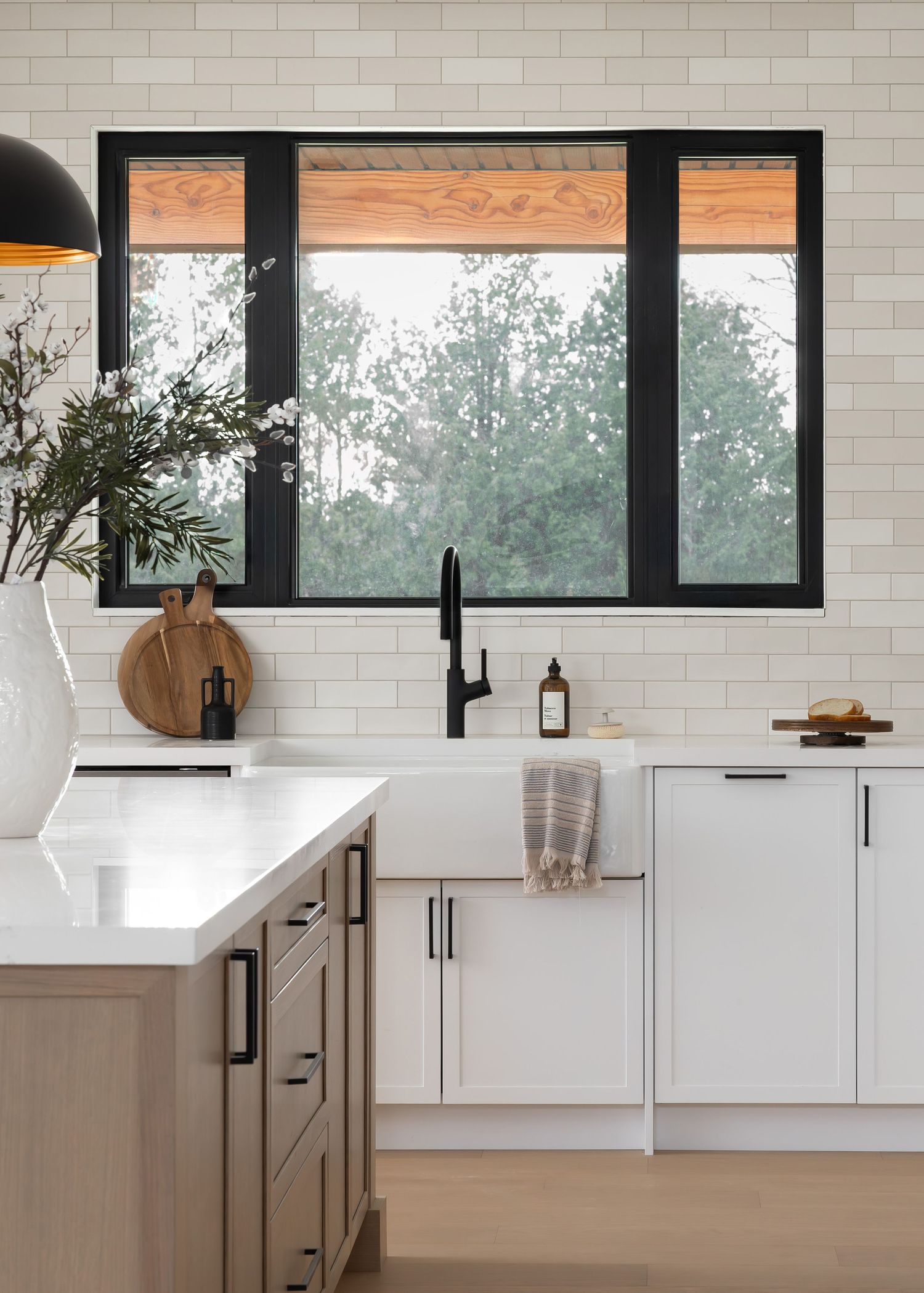 Modern kitchen with white cabinets, black-framed windows, and subway tile backsplash overlooking trees.
