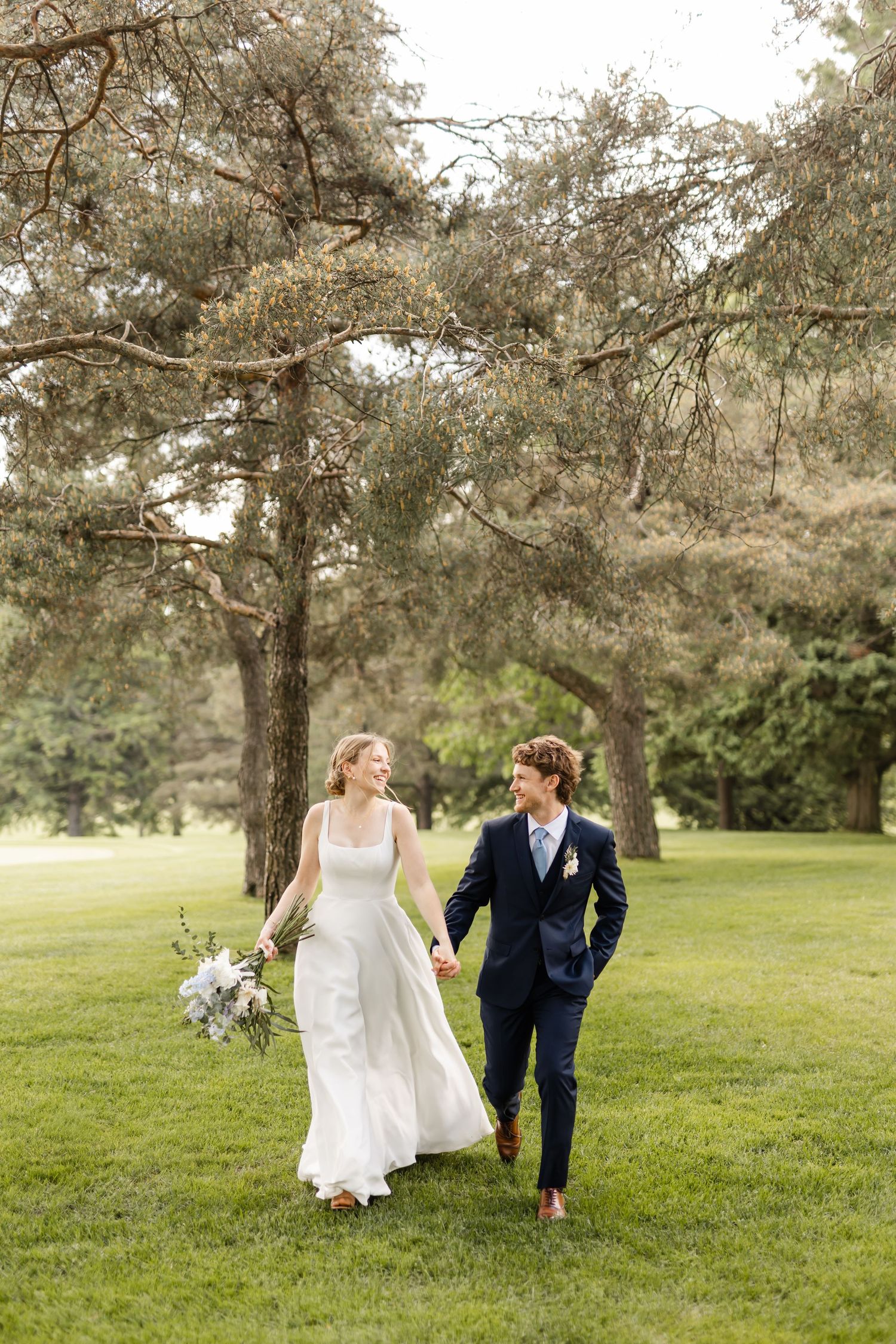 Bride in flowing white dress and groom in navy suit walking through scenic park.