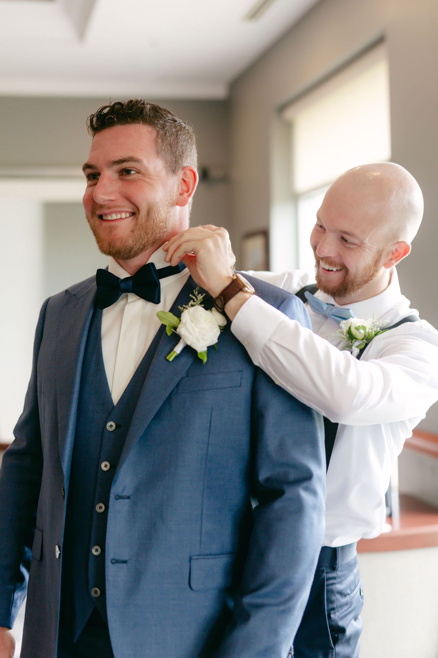 Groomsmen helping adjust a navy blue tuxedo and bowtie before the wedding ceremony.