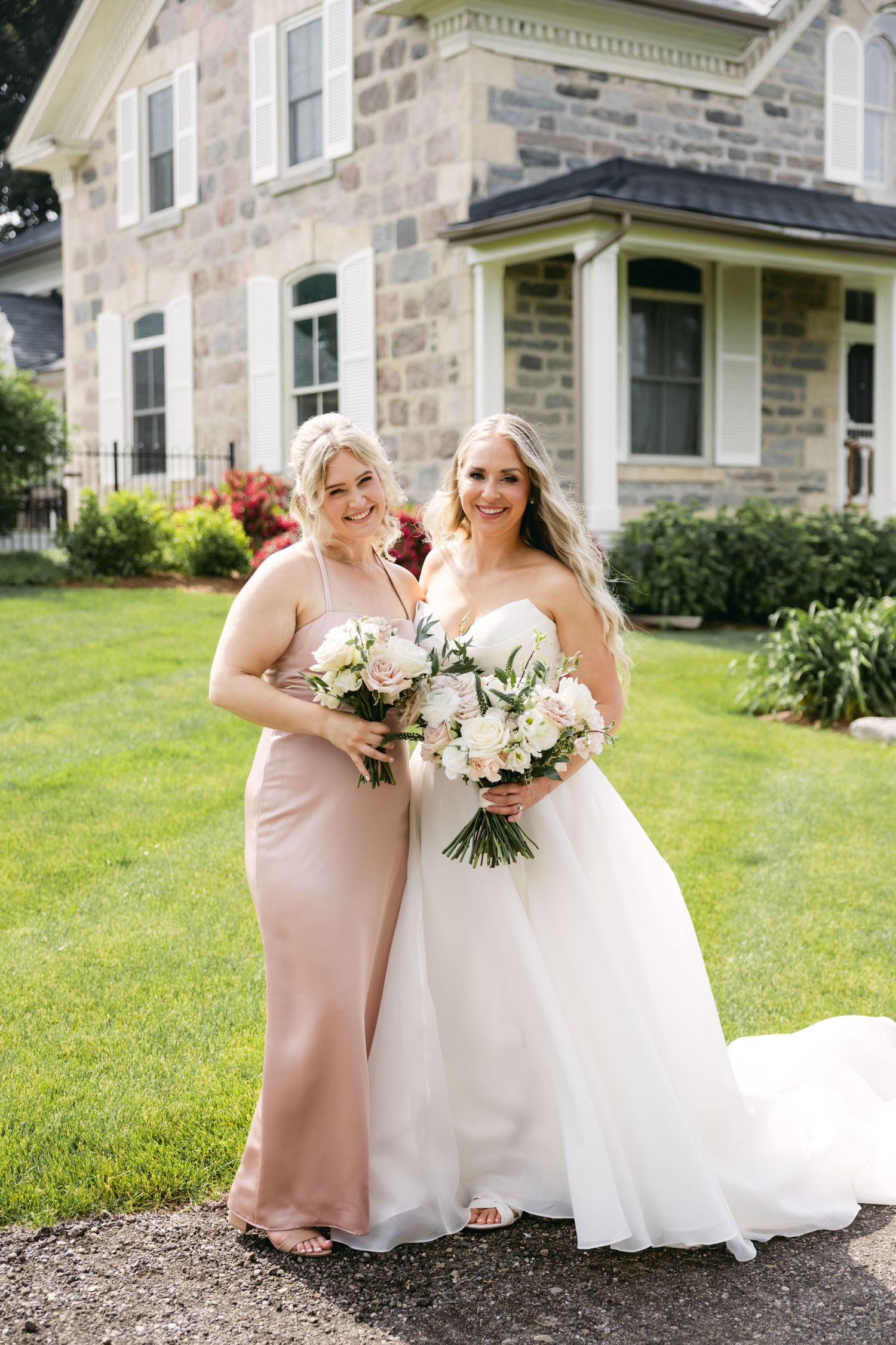 Wedding party members in pink and white dresses pose together on sunny lawn.