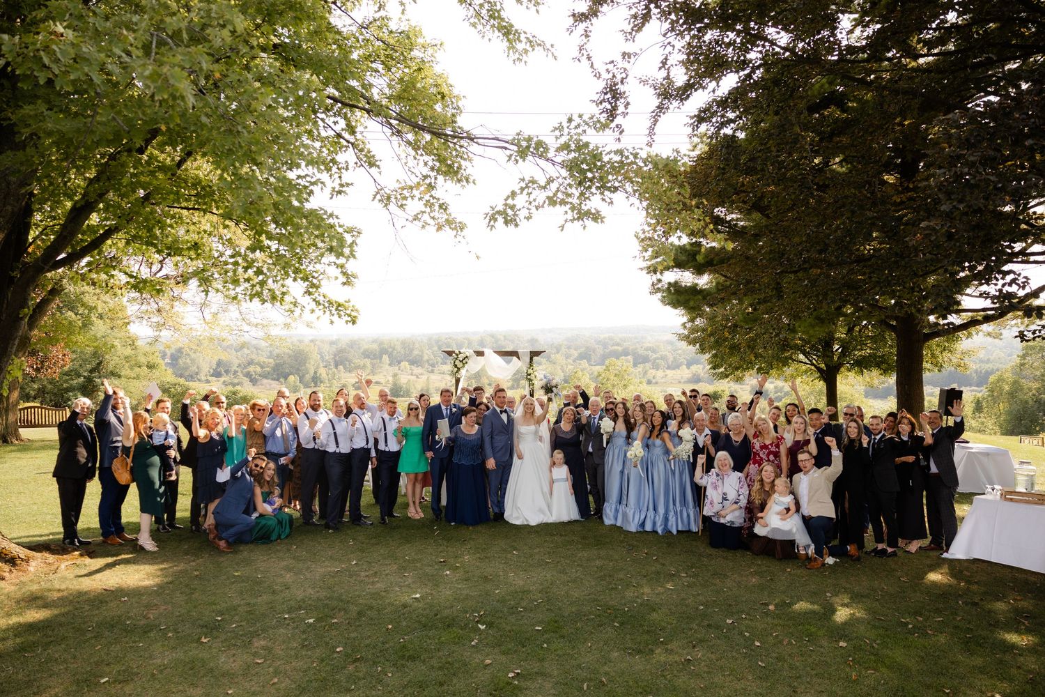 Large wedding group photo with guests gathered outdoors under trees at sunset.