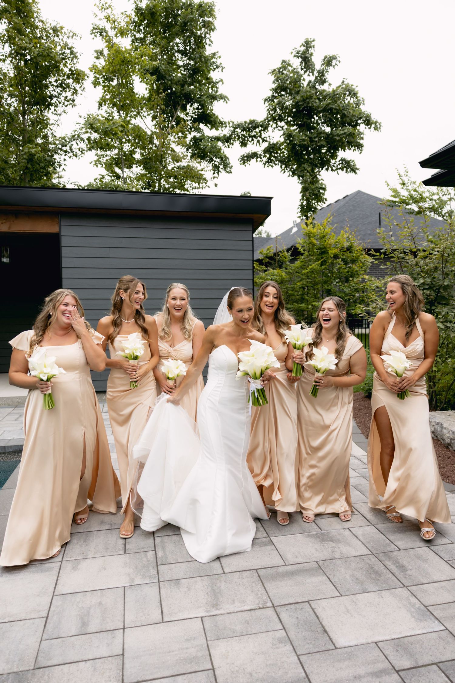 Bride in white gown laughing with six bridesmaids in champagne satin dresses holding white bouquets outdoors.