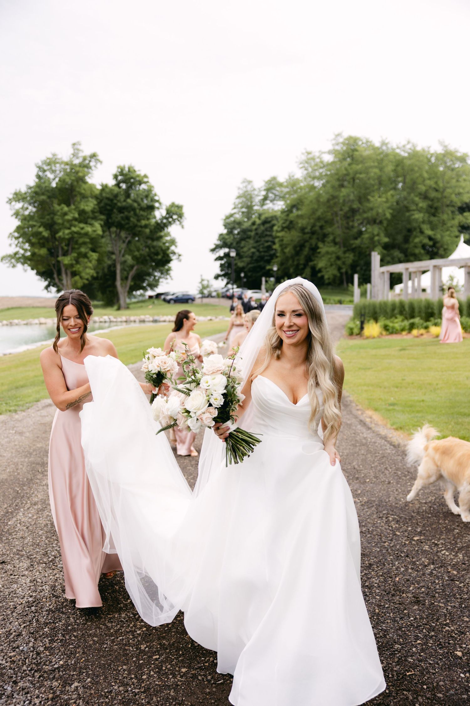 Bridal party in pink dresses walk along gravel path on sunny wedding day.
