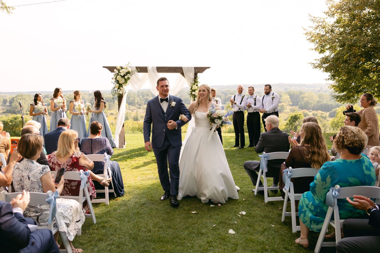 Wide angle view of wedding ceremony with guests seated on lawn chairs under trees.