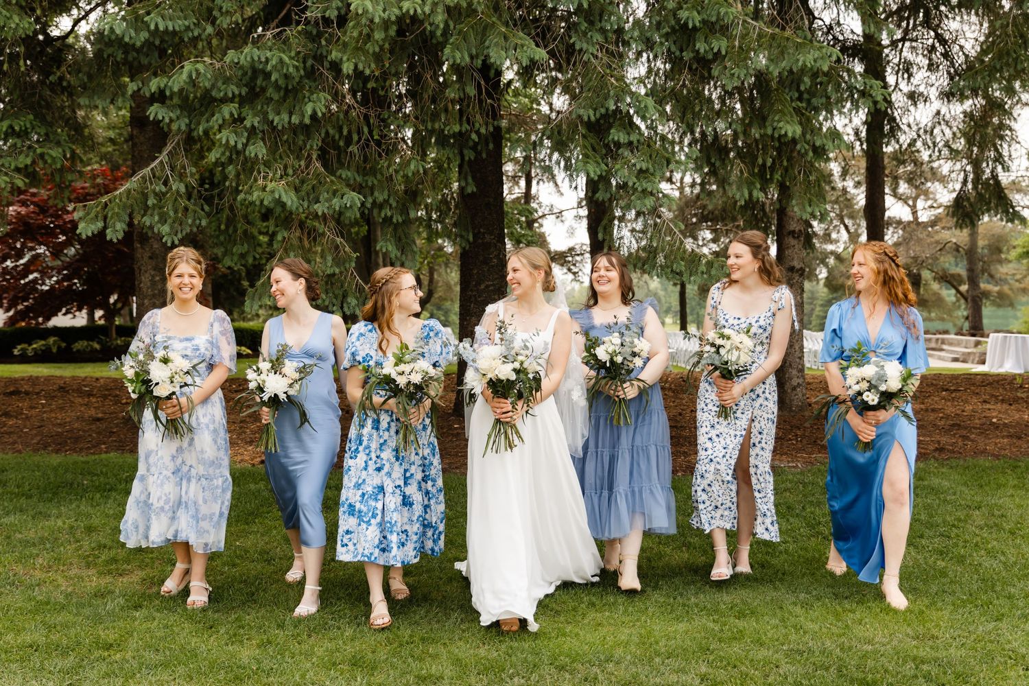 Wedding party in light blue dresses sits together on wooden chairs outdoors under evergreen trees.