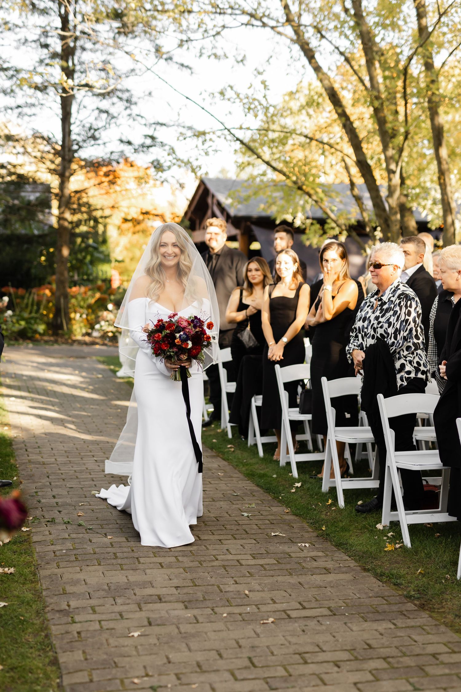 Wedding ceremony processional on stone pathway with white chairs and autumn foliage.