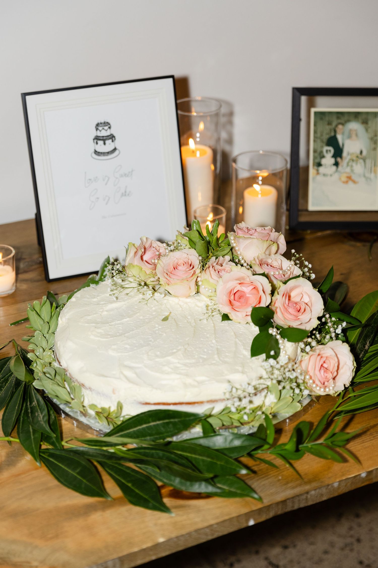 White wedding cake decorated with pink roses and greenery on a rustic wooden table.