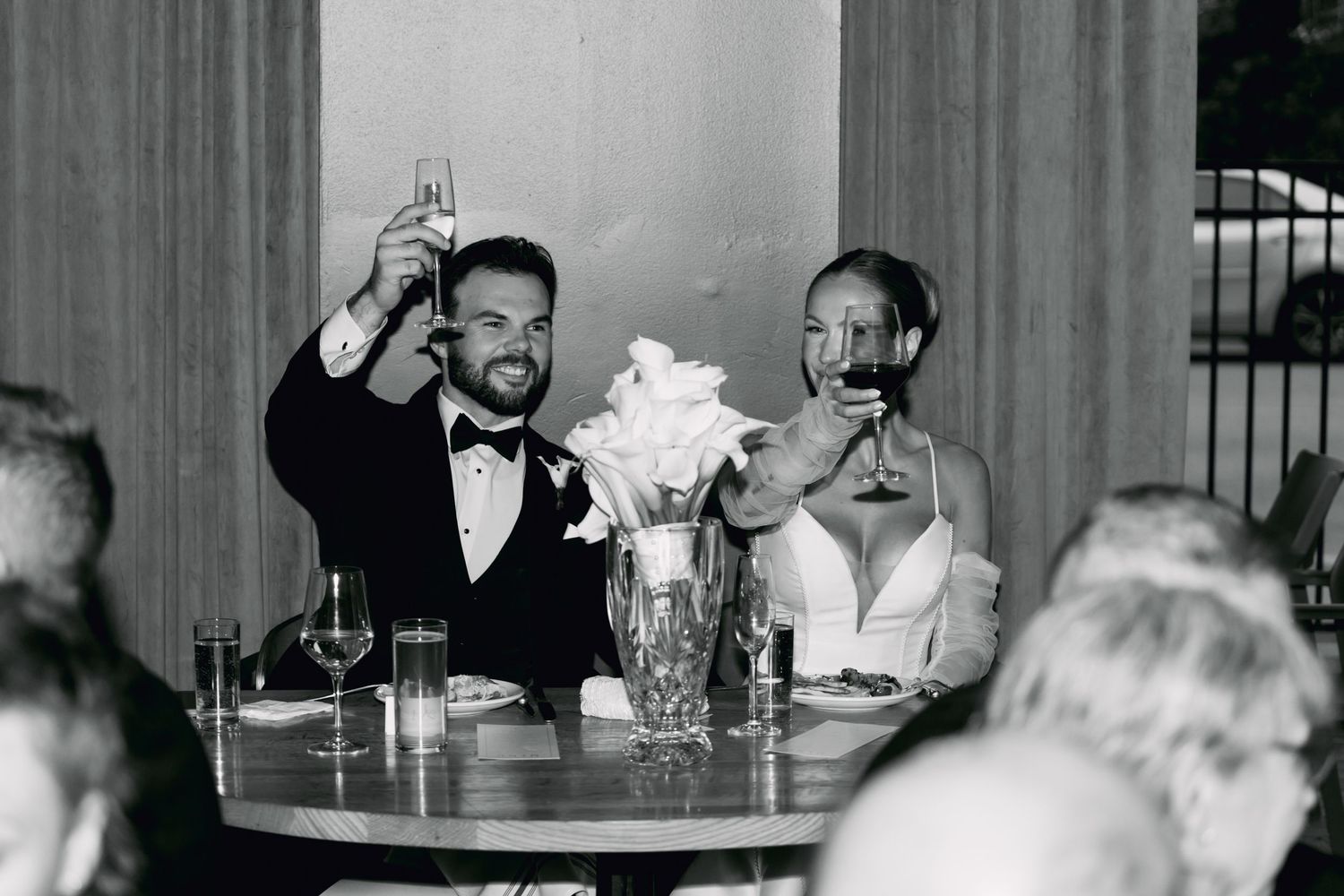 Black and white photo of newlyweds toasting champagne at wedding reception dinner table.