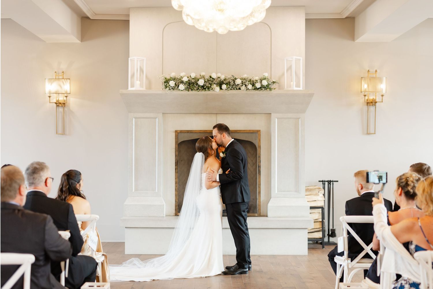 Bride and groom share their first kiss at the altar before seated wedding guests in a bright venue.