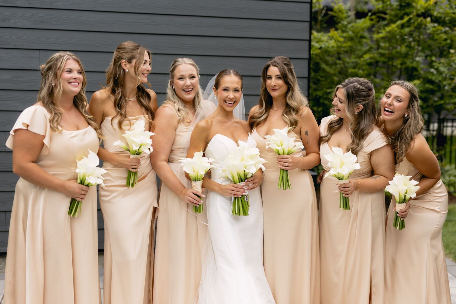 Bride in white strapless gown laughs with six bridesmaids in matching champagne dresses holding white bouquets.