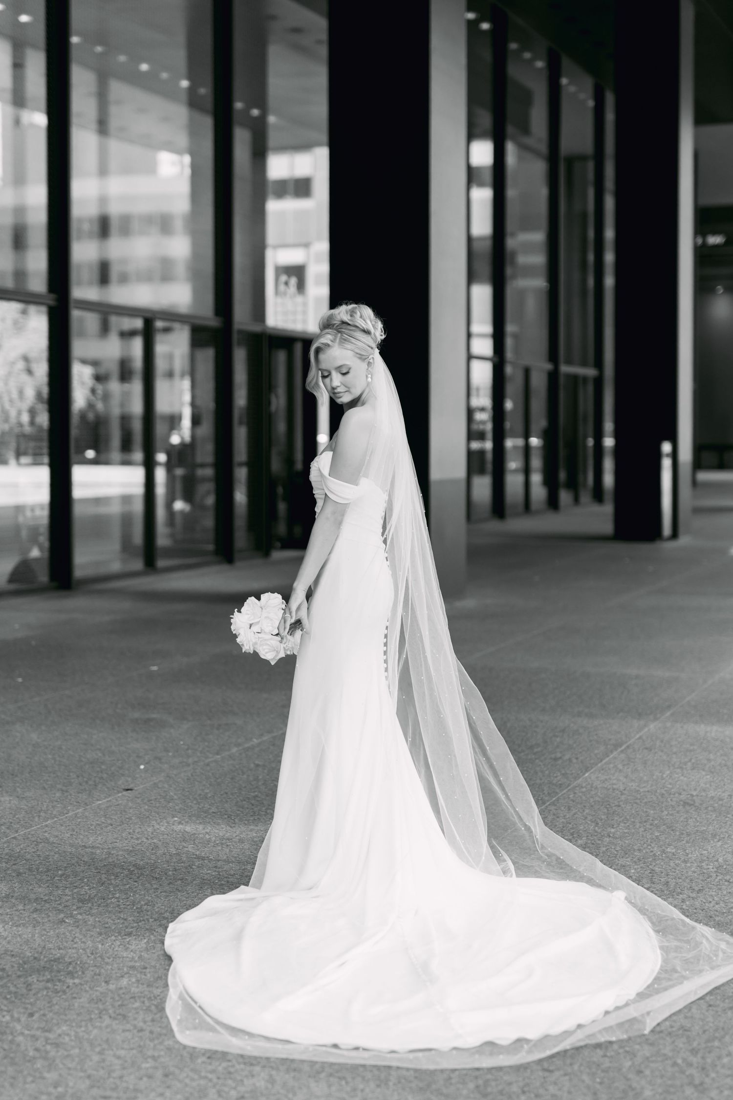 Black and white portrait of bride in off-shoulder gown with cathedral veil holding bouquet outside modern building.