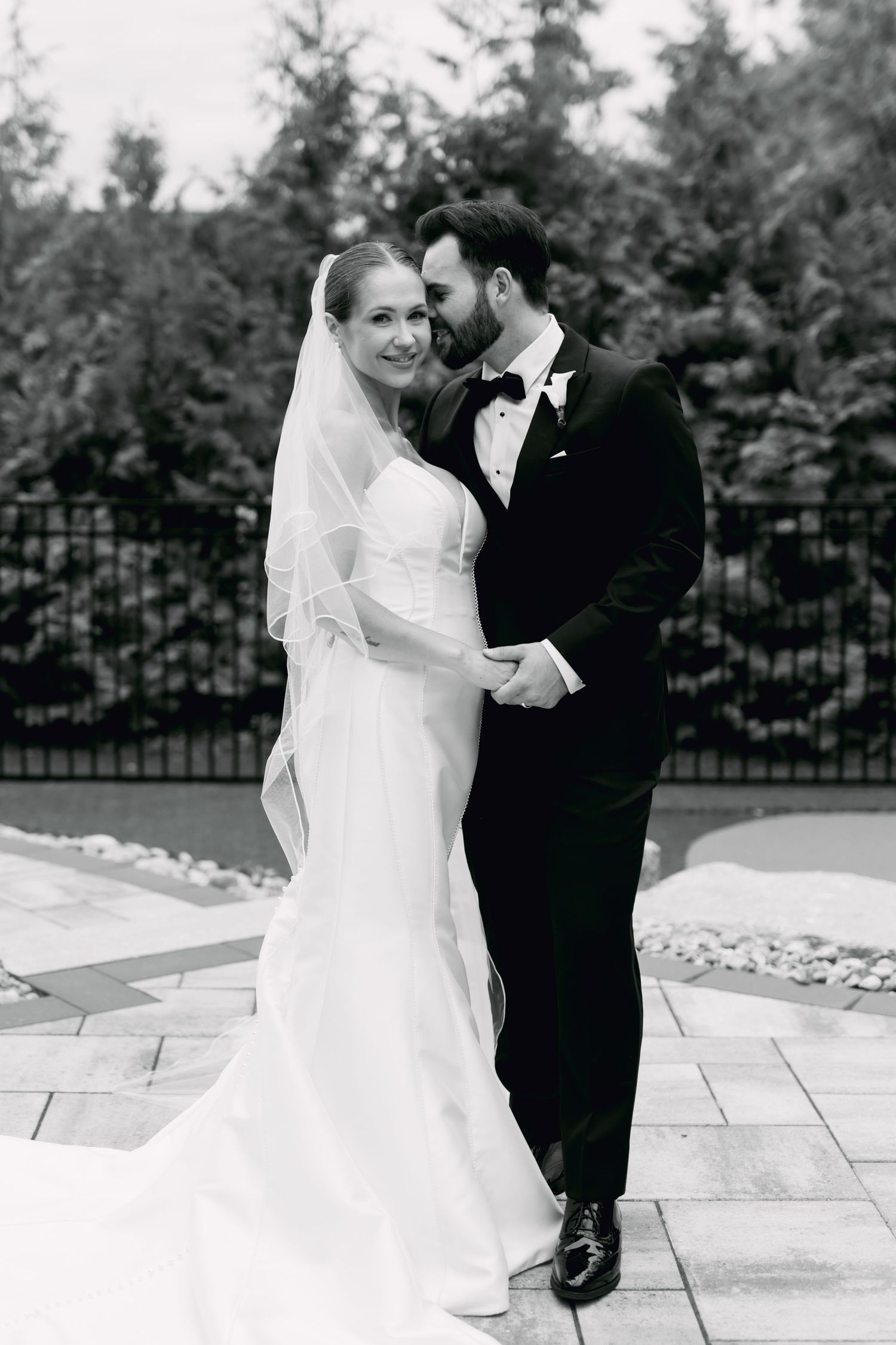 Black and white portrait of bride and groom embracing outdoors near iron fence and trees.