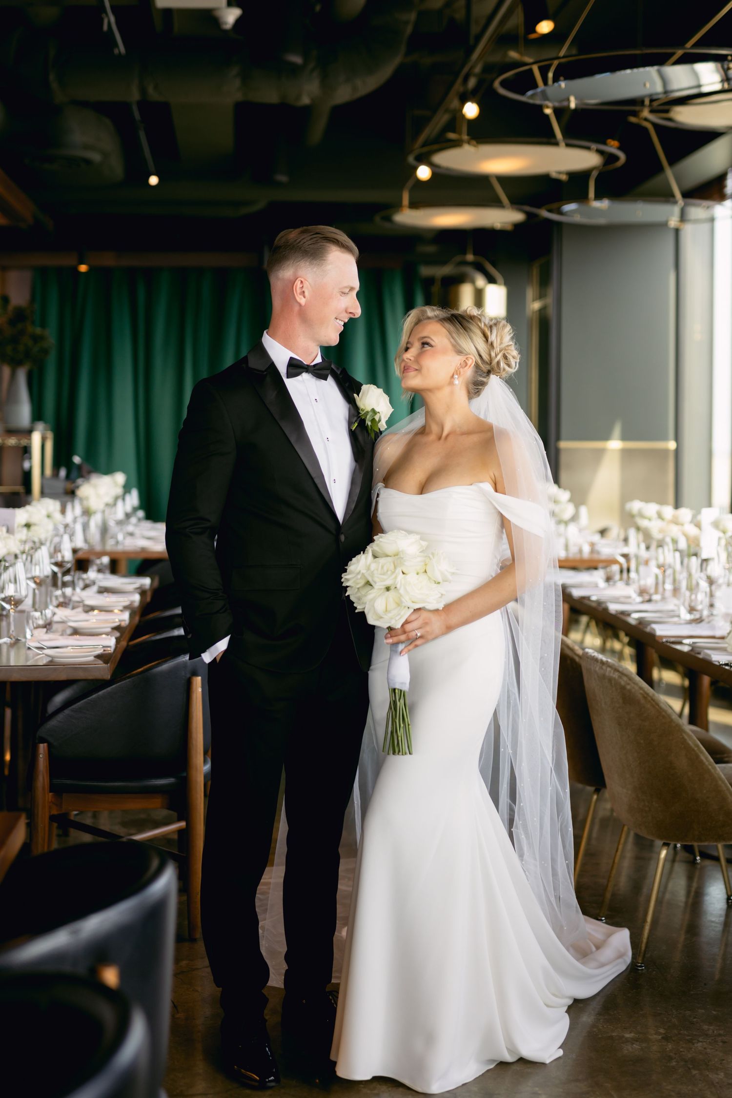 Bride holding white bouquet and groom in tuxedo pose in elegantly set wedding reception venue.