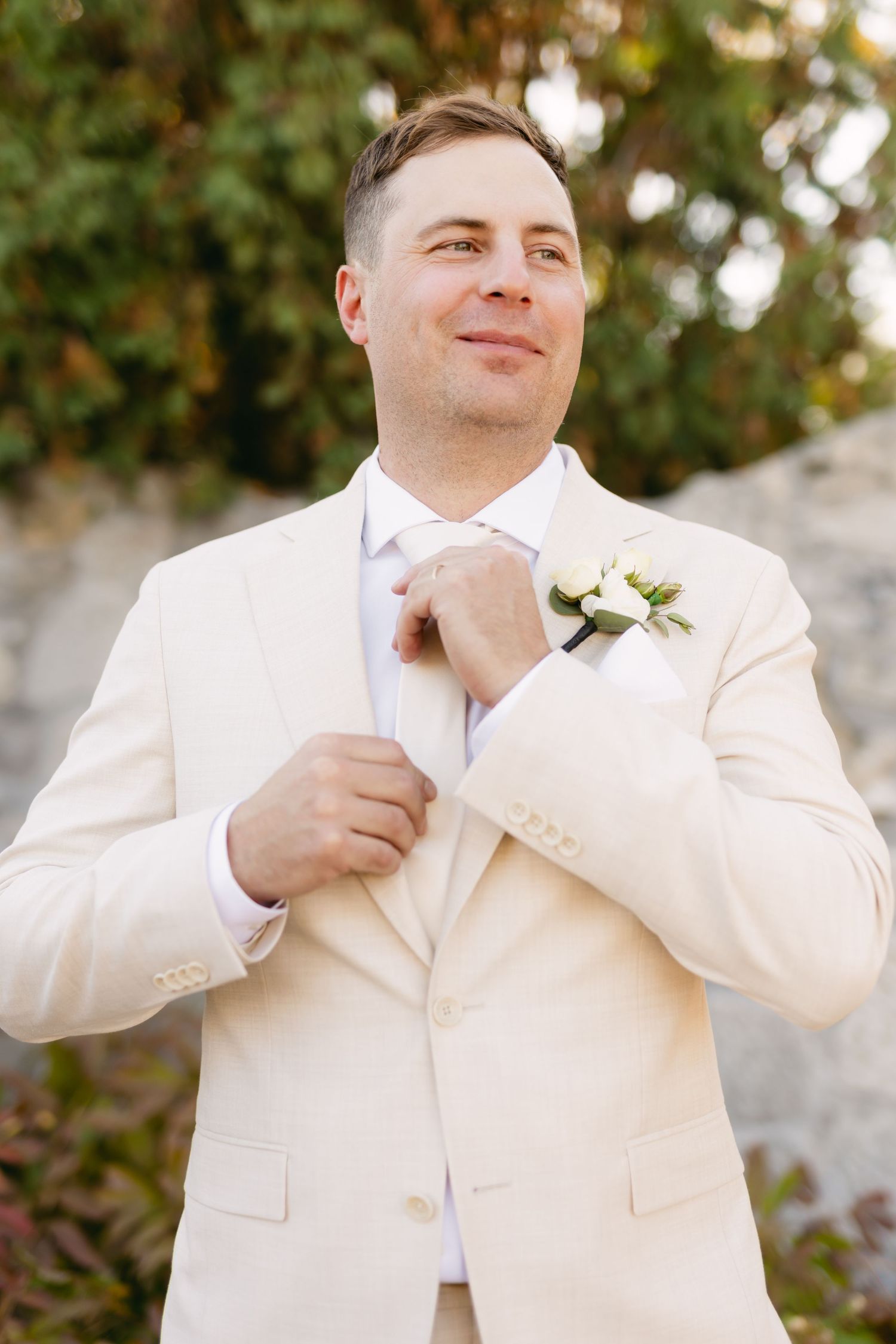A person in a cream suit smiles while adjusting their collar outdoors.