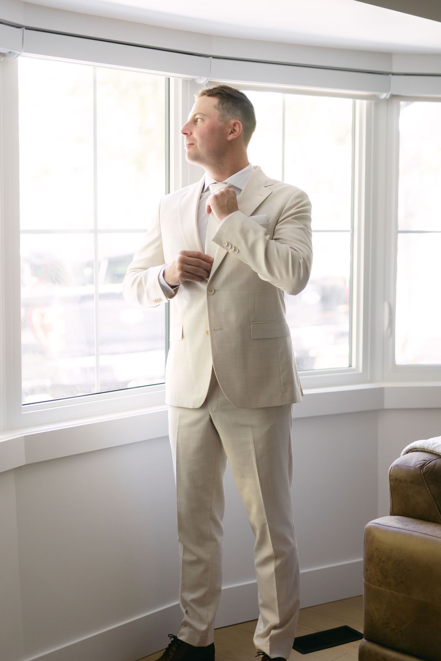 A person in a light colored suit poses in a bright room with large windows.