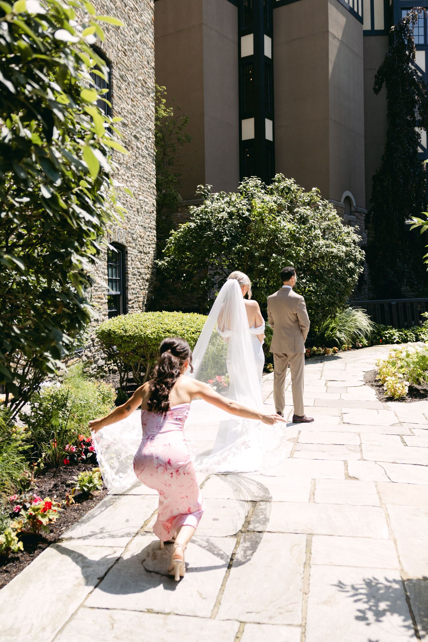 Wedding party member in pink dress helps with bride's veil on stone pathway.