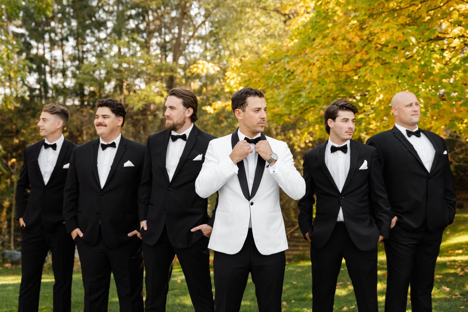 Groomsmen in tuxedos pose together against autumn trees.