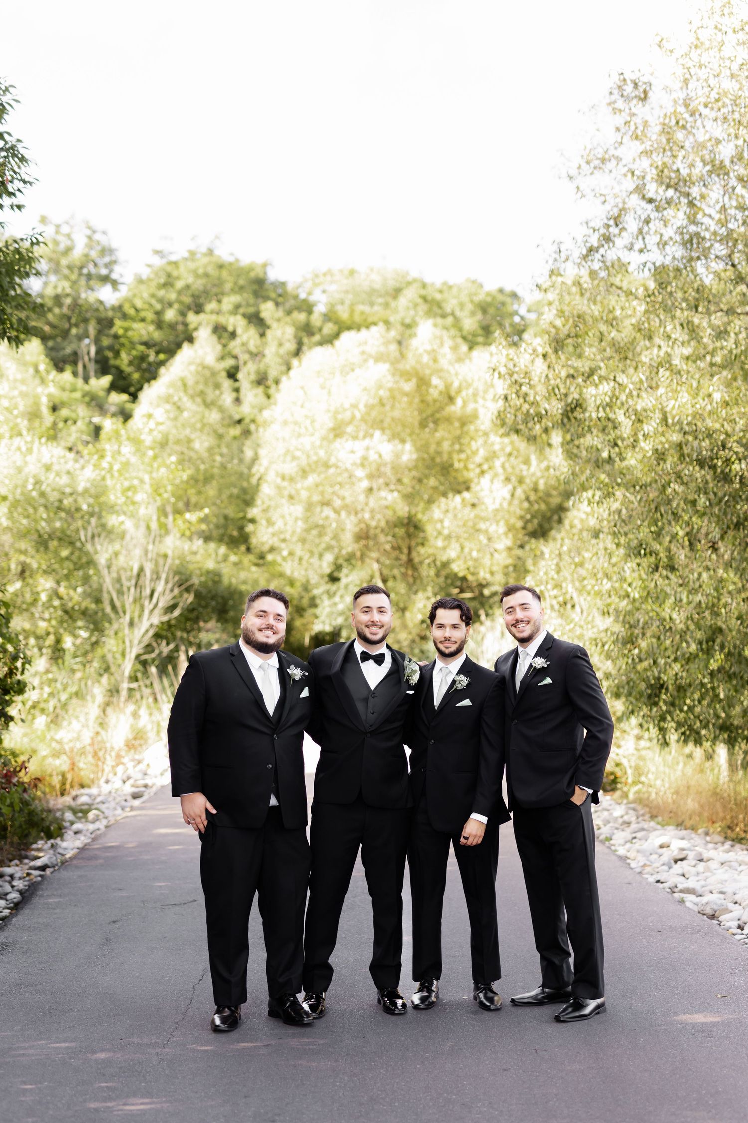 Wedding party in black tuxedos walking together on paved pathway surrounded by greenery.