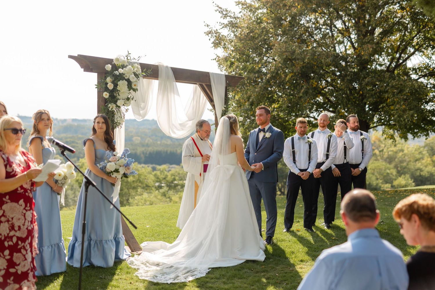 Wedding party in light blue and navy attire standing together under trees during an outdoor ceremony.