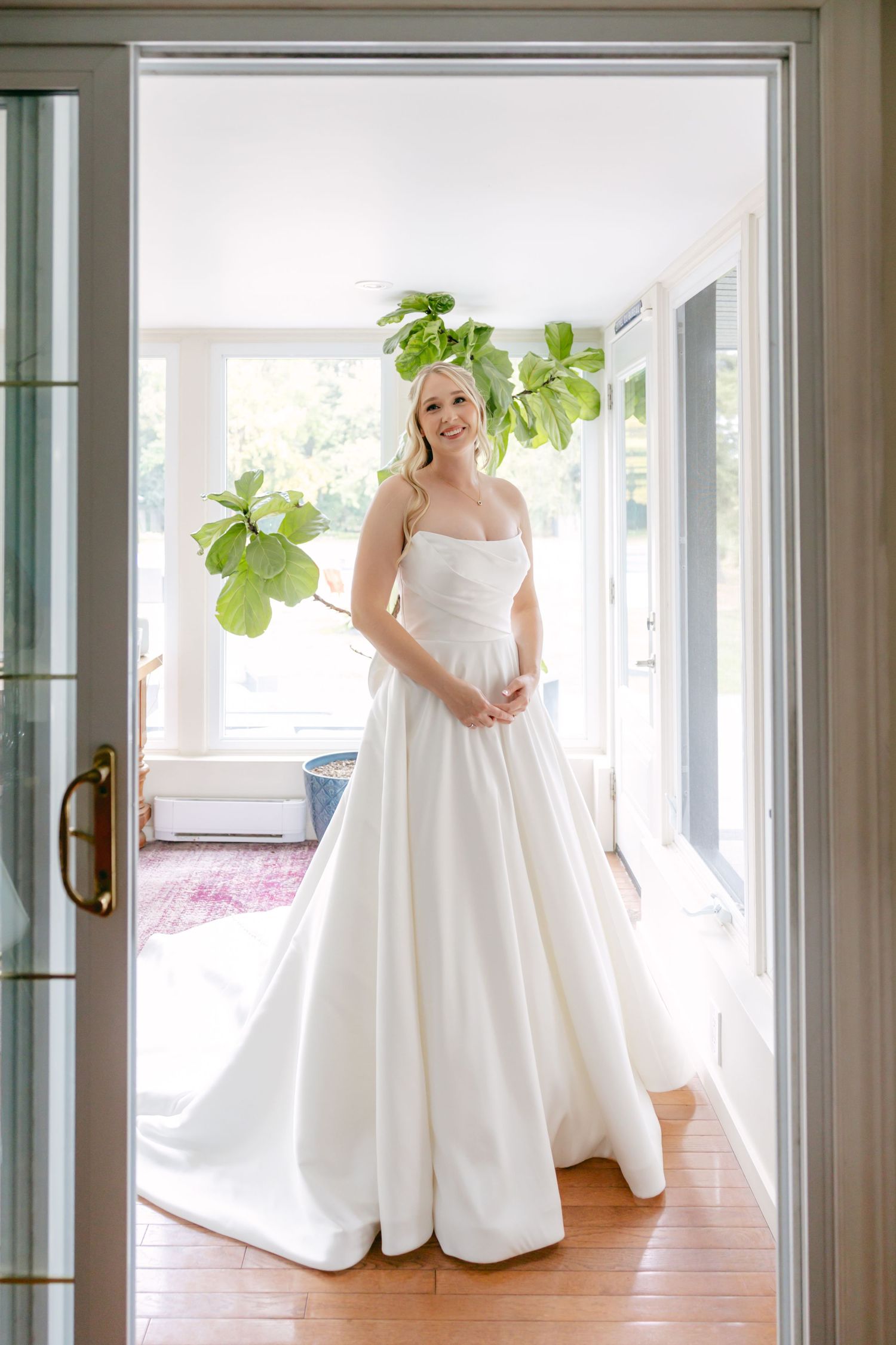 Bride in an elegant white ballgown poses in a bright room with floor-to-ceiling windows.