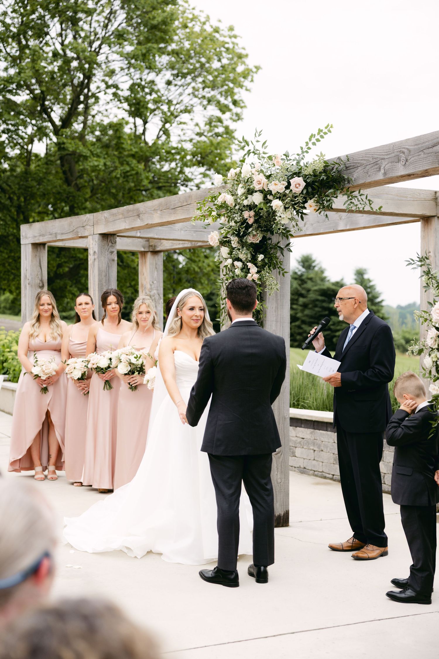 Wedding party in blush dresses and black suits gathered under a decorated wooden pergola.