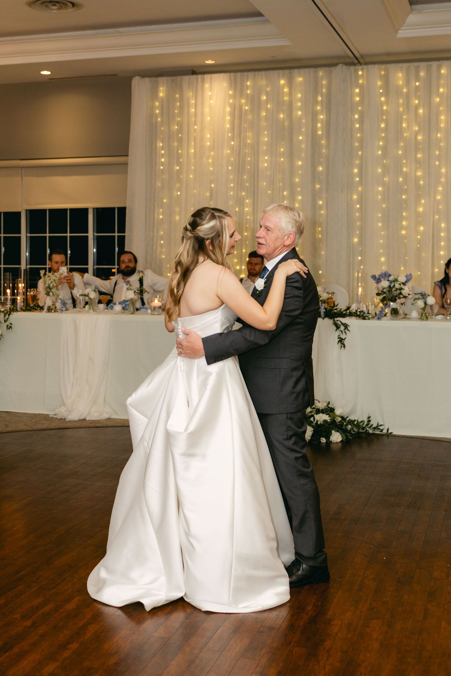 Elegant ballroom dance between partners in formal attire on hardwood floor with twinkle light backdrop.