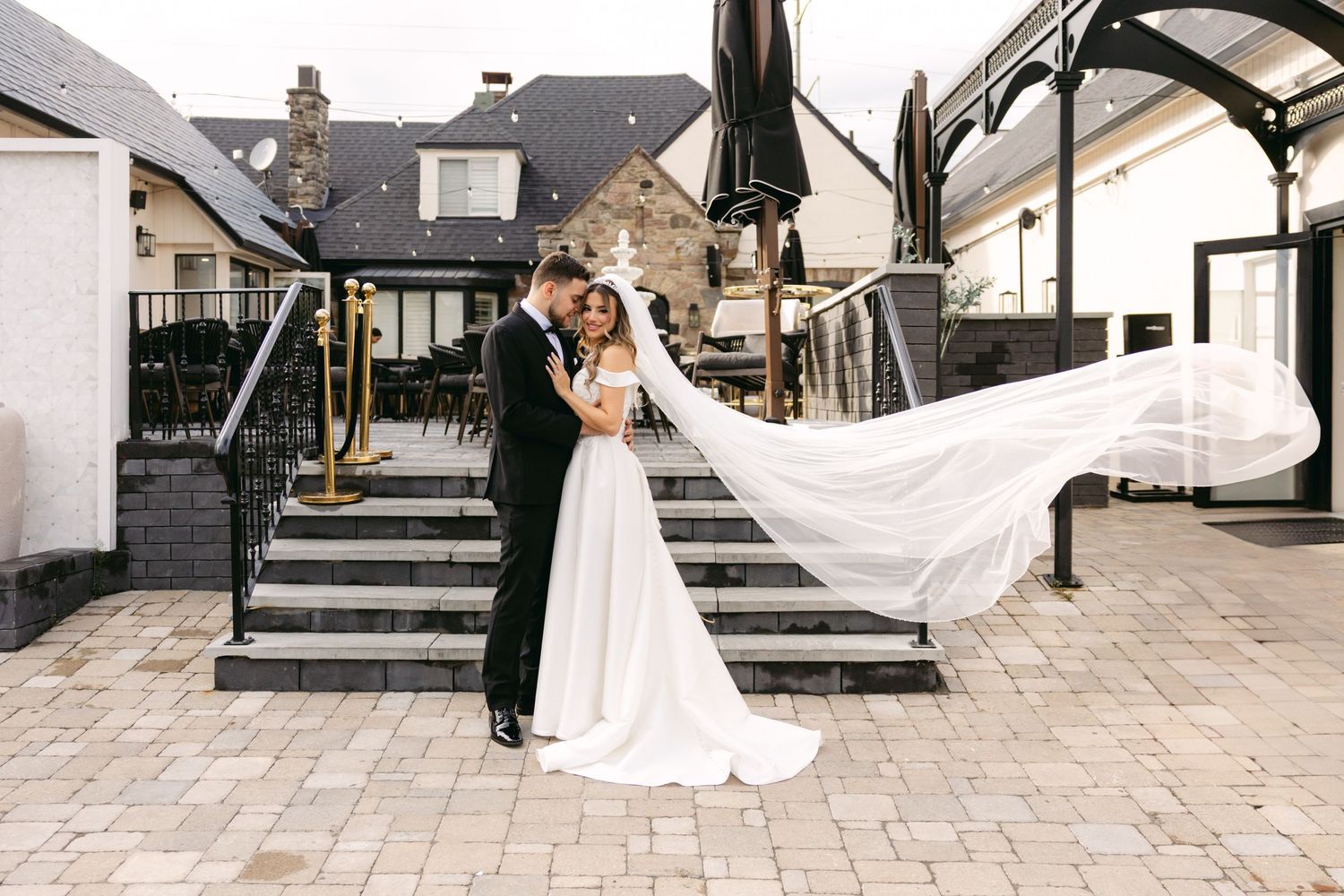 Wedding dress train and veil flow dramatically across stone patio during outdoor photo session.