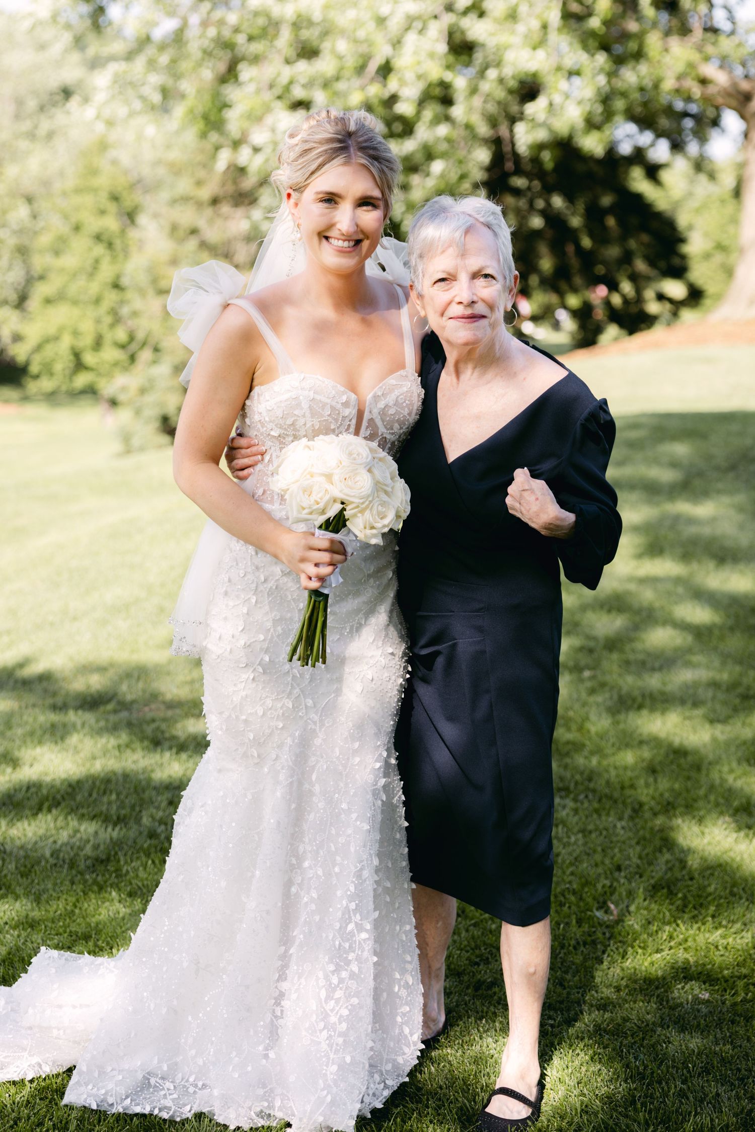 Bride in detailed lace gown posing with wedding party member on sunny lawn.