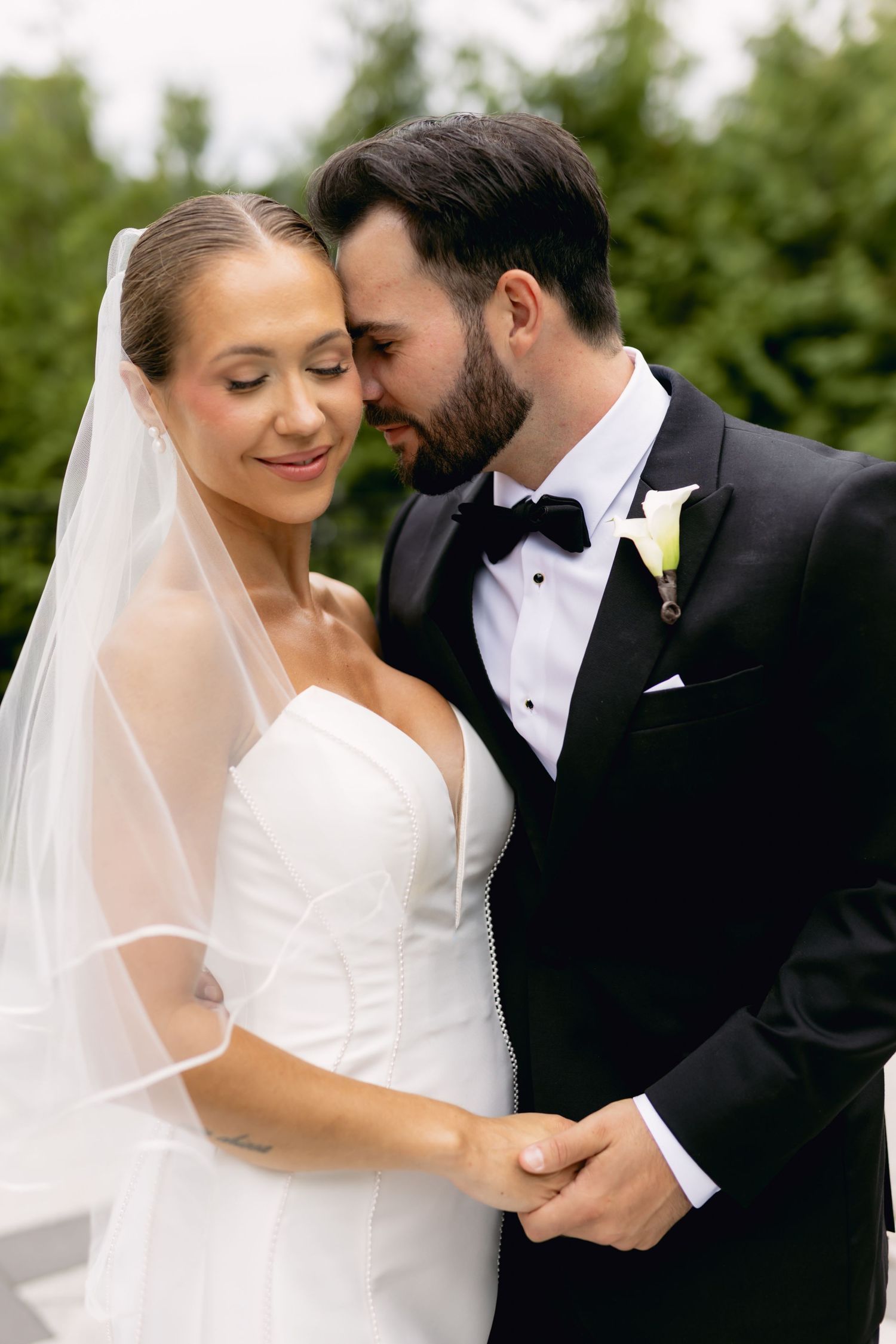 Groom in black tuxedo tenderly holds bride in white strapless gown with veil outdoors.